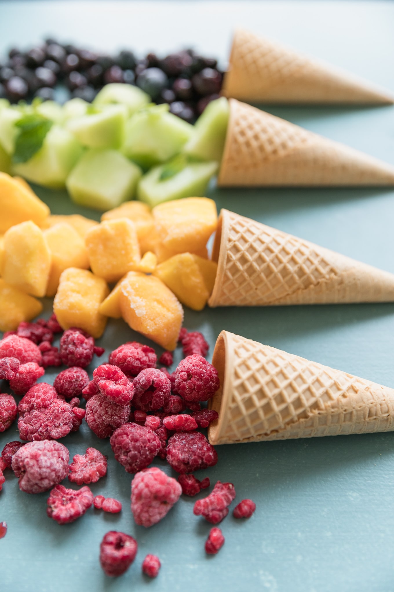 Close up shot of four ice cream cones with raspberries, mango, honeydew, and blueberries pouring out of them