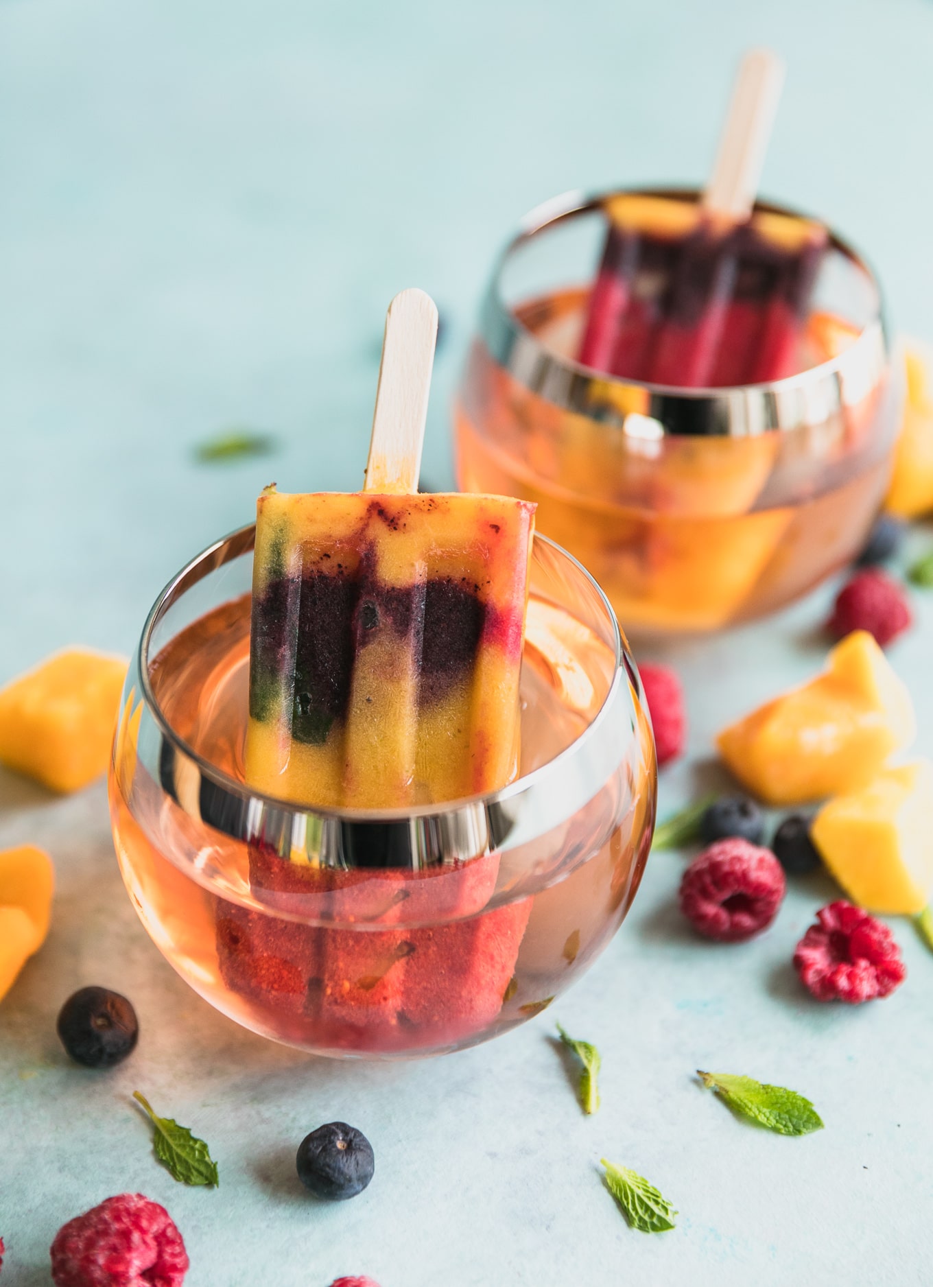 Close up shot of two glasses of rosé with a rainbow popsicle sitting in it and fruit scattered around
