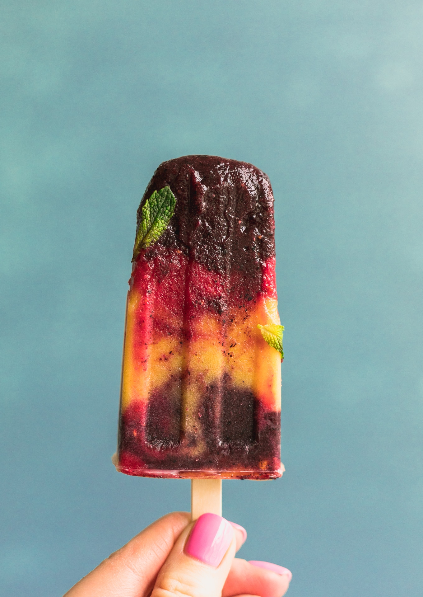 Close up forward facing shot of a layered blueberry, raspberry and mango popsicle with a mint leaf on it against a blue background