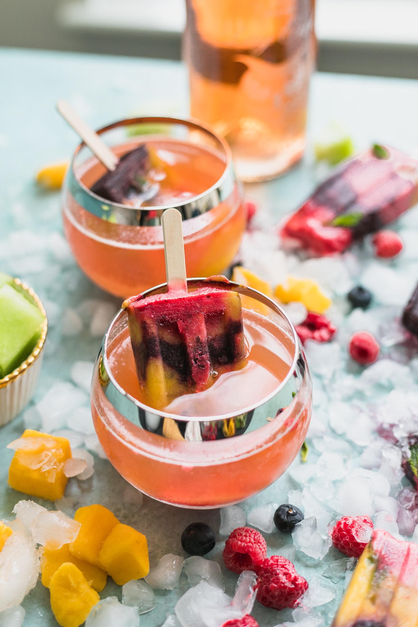 Close up shot of two glasses of rosé with a rainbow popsicle sitting in it and fruit scattered around