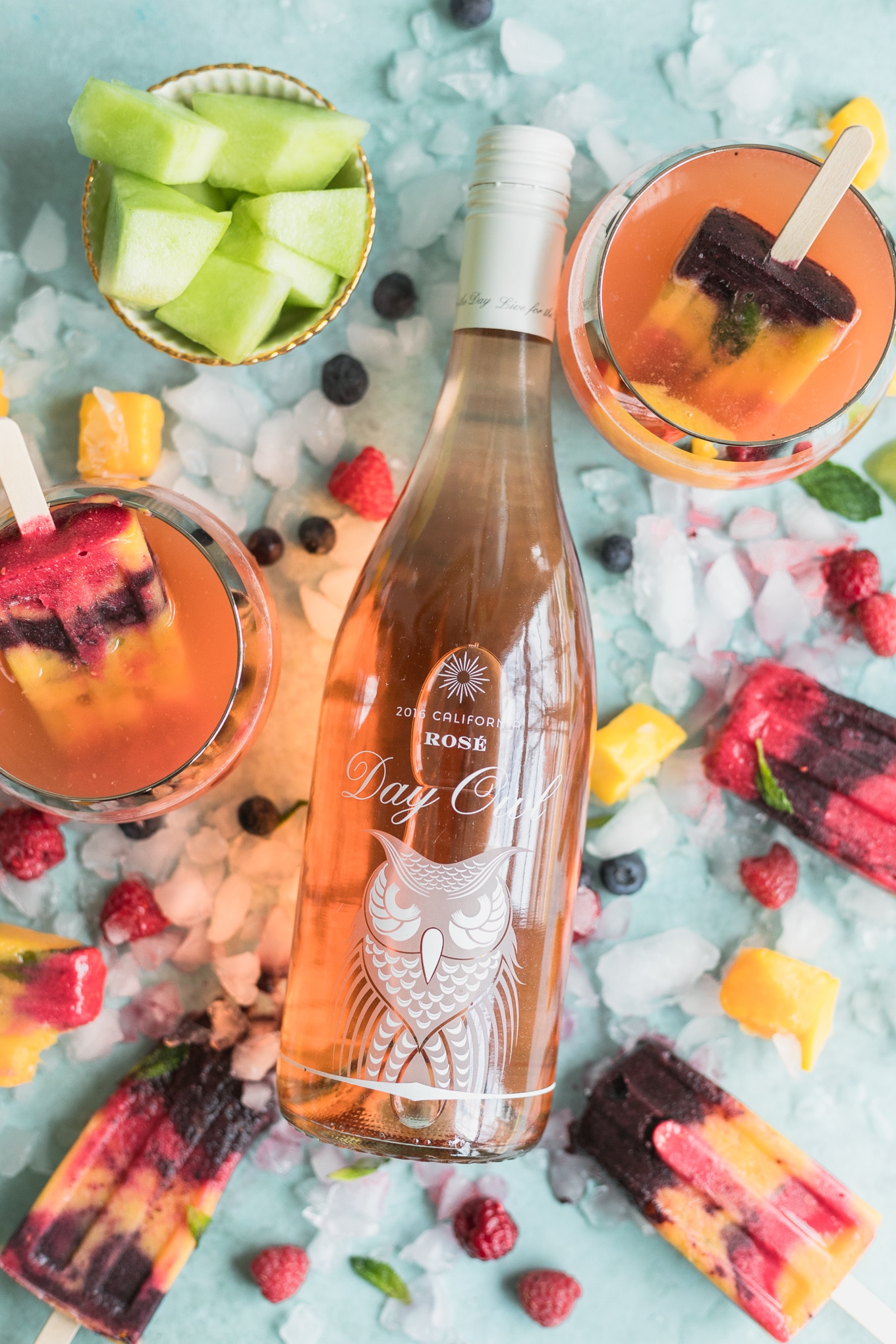 Overhead shot of a bottle of rosé surrounded by glasses of rosé with popsicles in them, crushed ice, fruit, and rainbow popsicles