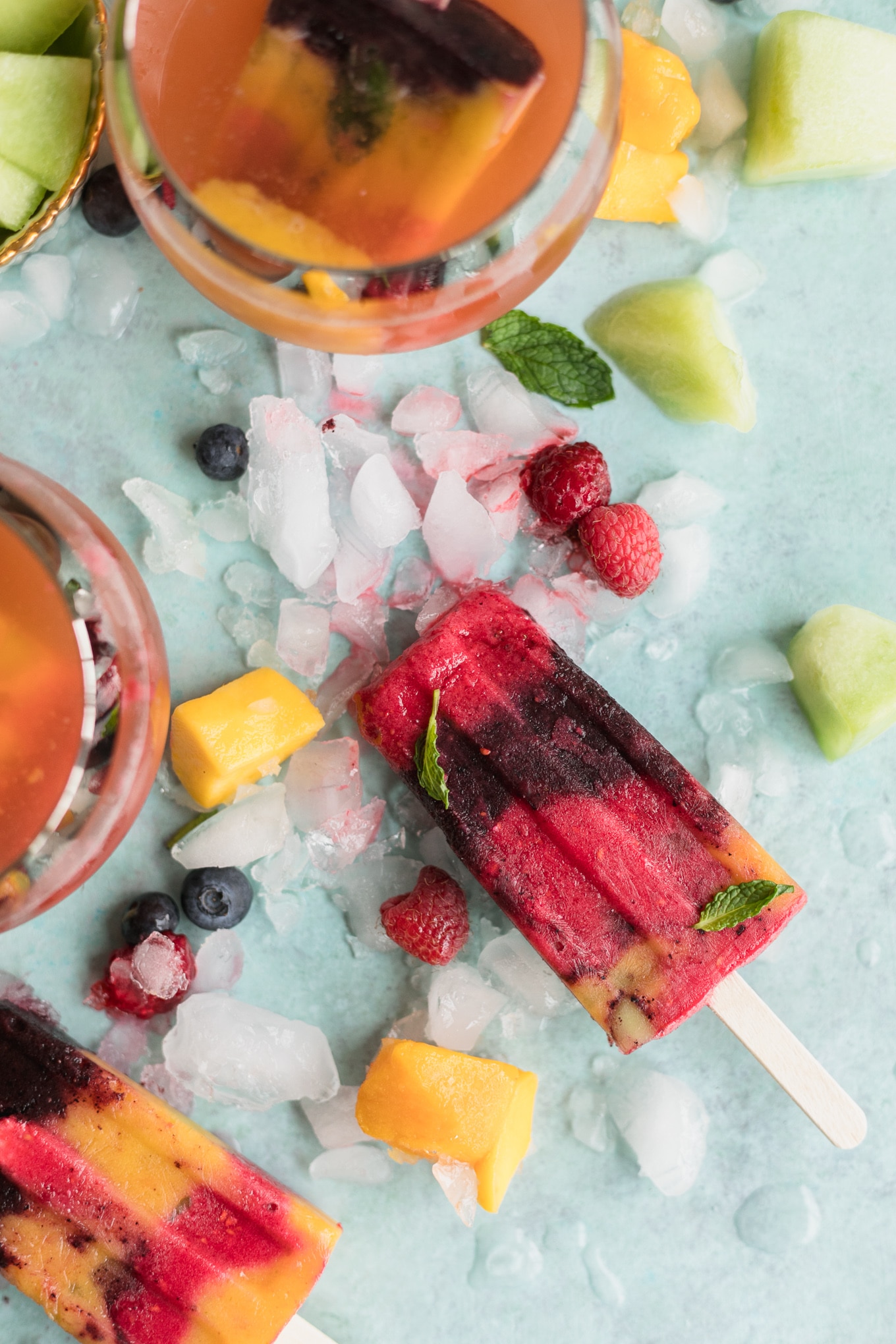 Overhead shot of a raspberry and blueberry popsicle on an aqua background with ice and fruit scattered around, and two glasses of rosé next to it
