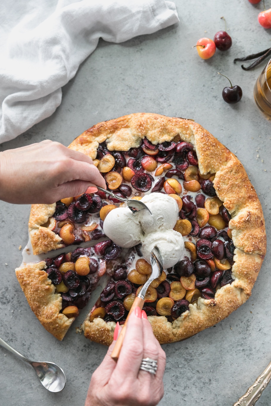 Overhead shot of a cherry galette with 3 scoops of ice cream on top with two hands reaching in with spoons to eat the ice cream