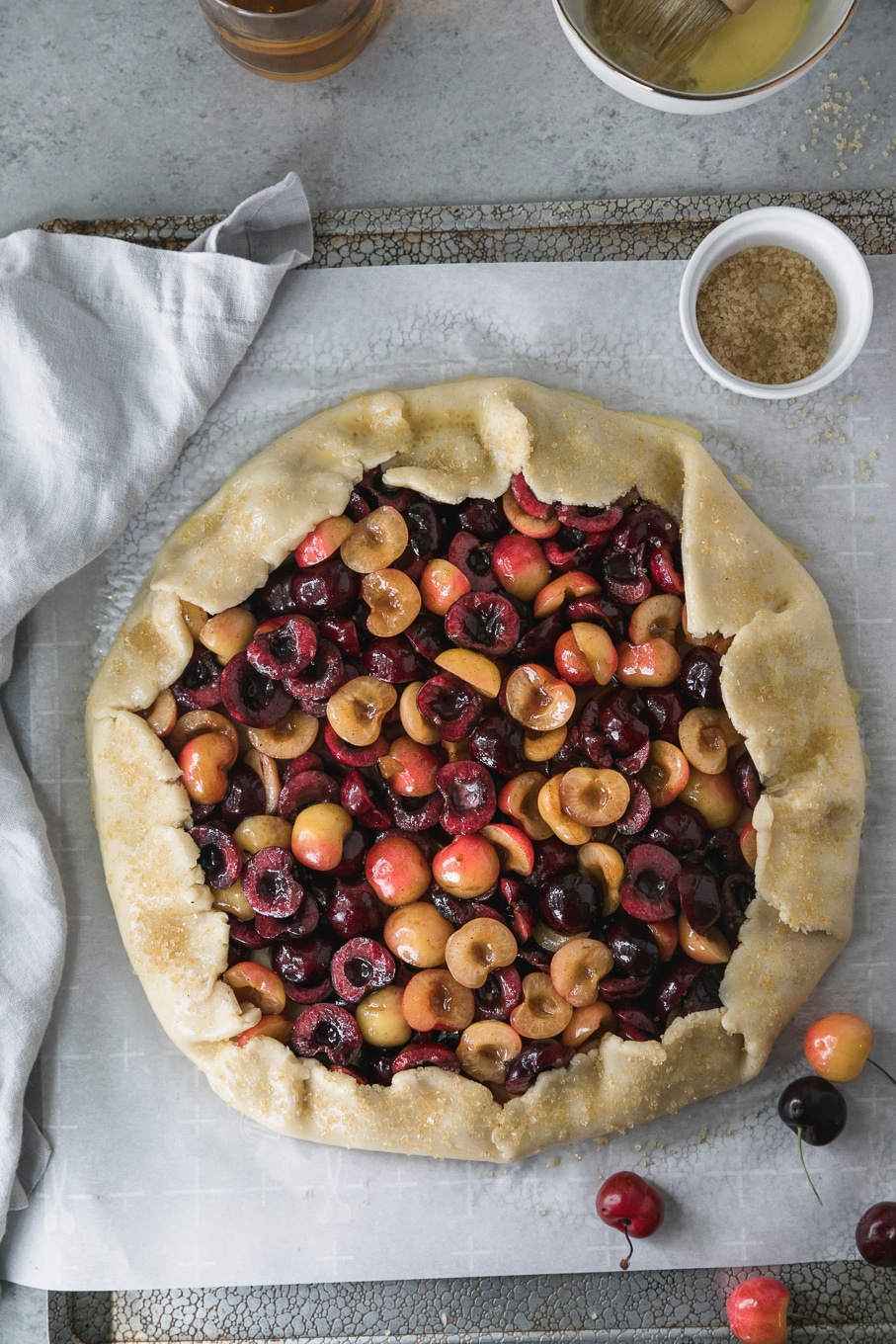 Overhead shot of a cherry galette on a baking sheet with a light grey napkin to the left