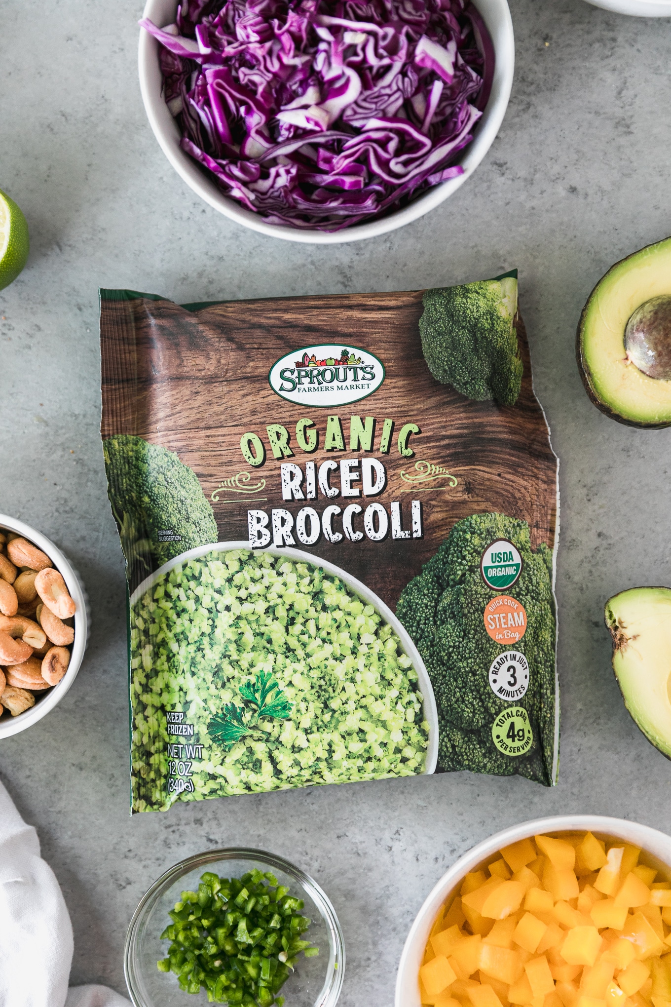 Overhead close up shot of a bag of riced broccoli surrounded by a halved avocado, bowl of red cabbage, bowl of diced bell pepper, and cashews