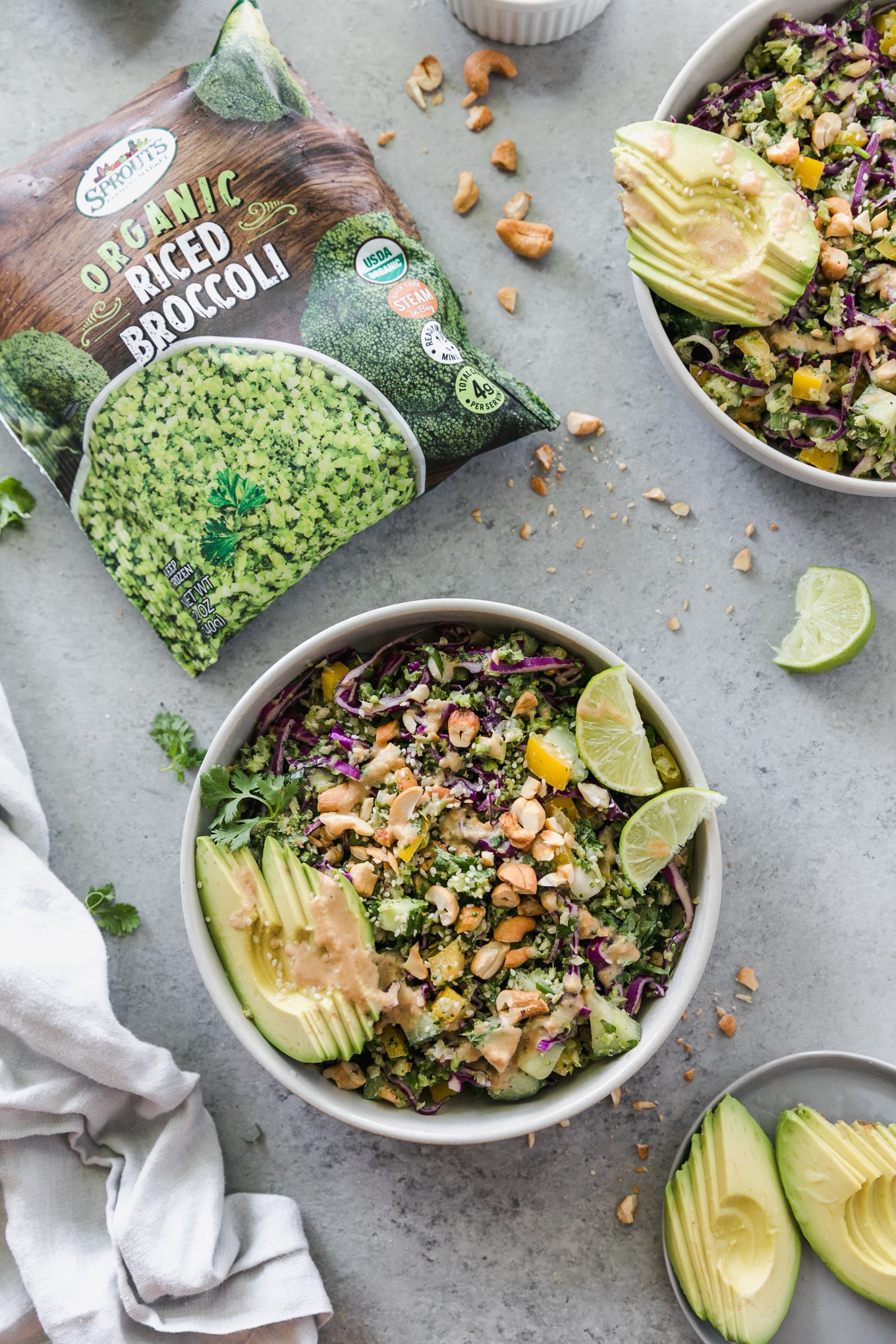Overhead shot of a salad topped with avocado, lime wedges, and a bag of riced broccoli