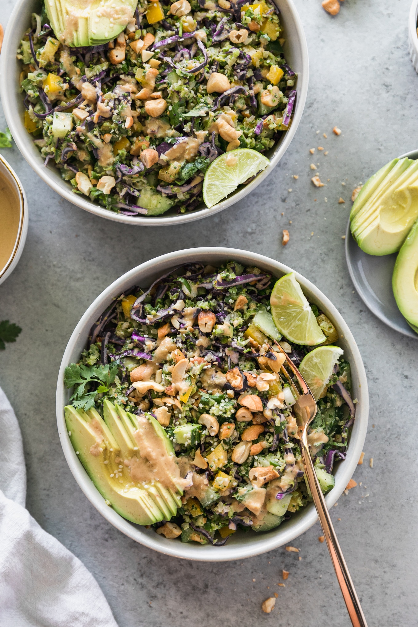 Overhead shot of a salad with cashews, avocado, lime wedges, and a gold fork sticking out of the bowl