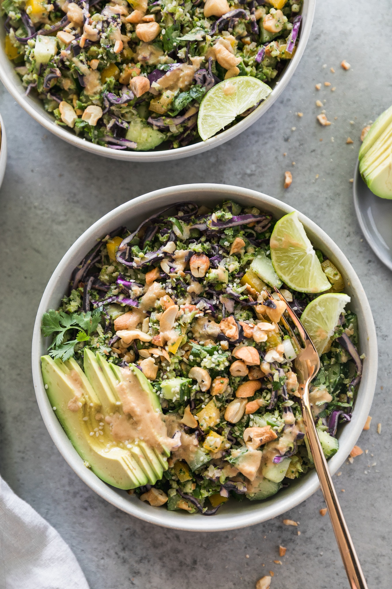 Overhead close up shot of a salad with cashews, avocado, lime wedges, and a gold fork sticking out of the bowl