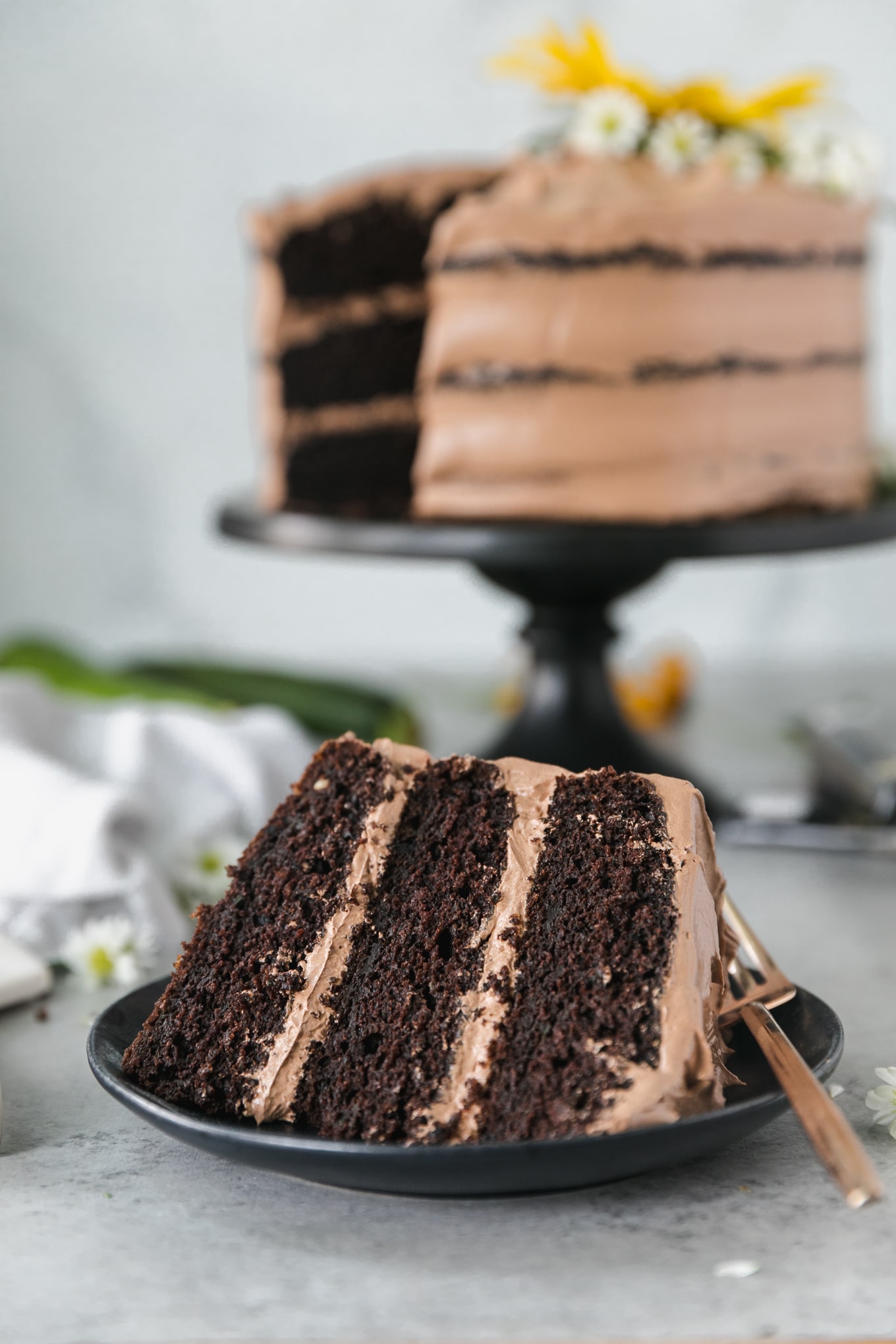 Close up shot of a piece of chocolate cake with the full later cake in the background on a black cake stand