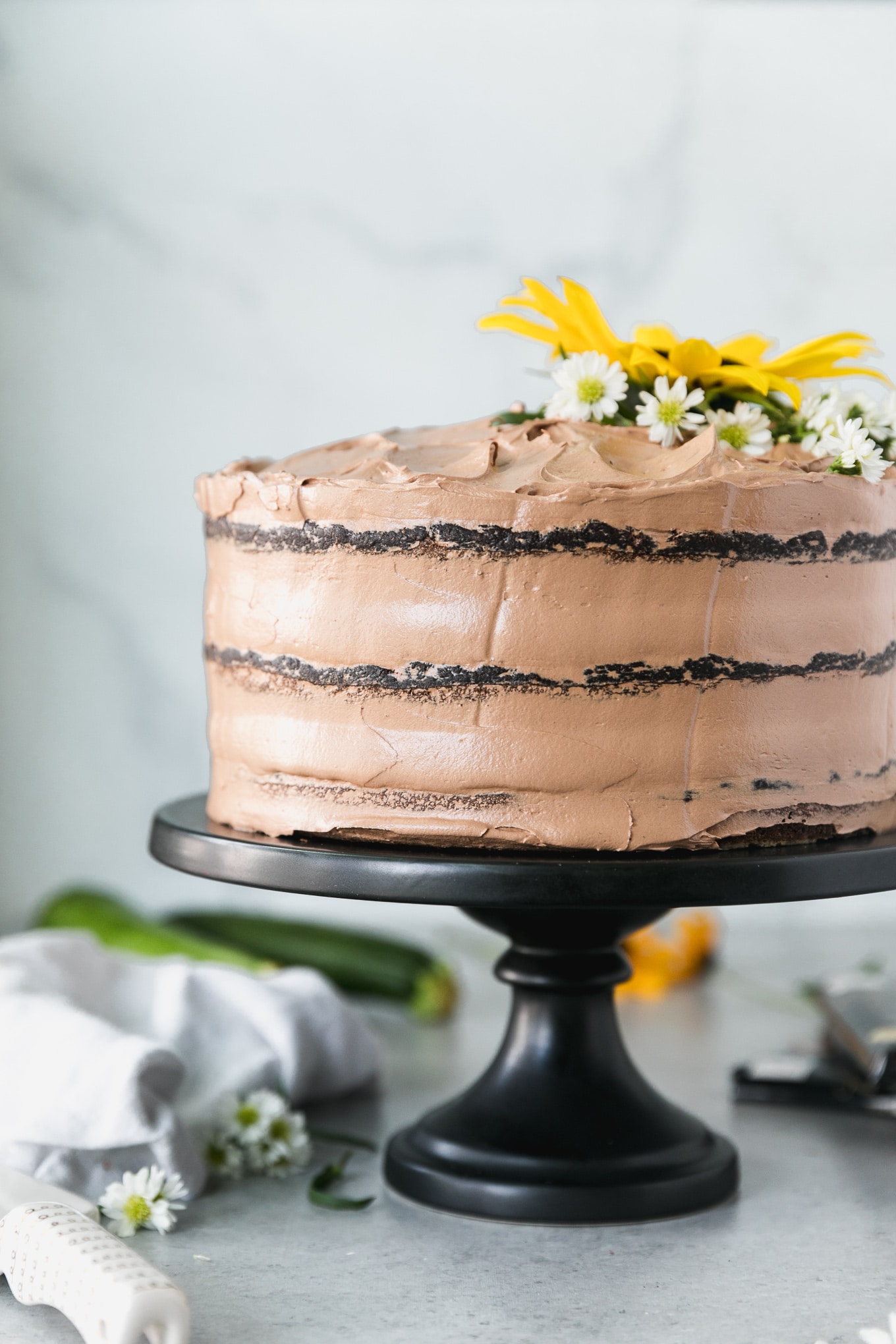 Forward facing shot of a 3 layer chocolate cake topped with a sunflower and small white flowers and a napkin and a zucchini in the background