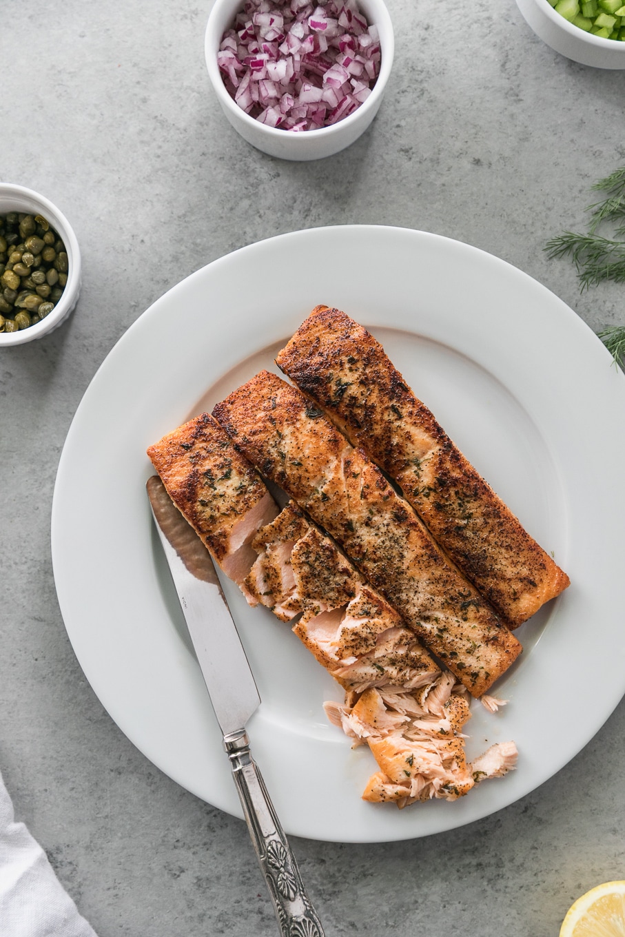 Overhead shot of a plate of 3 salmon fillets, a bowl of diced red onion, a bowl of capers, and dill