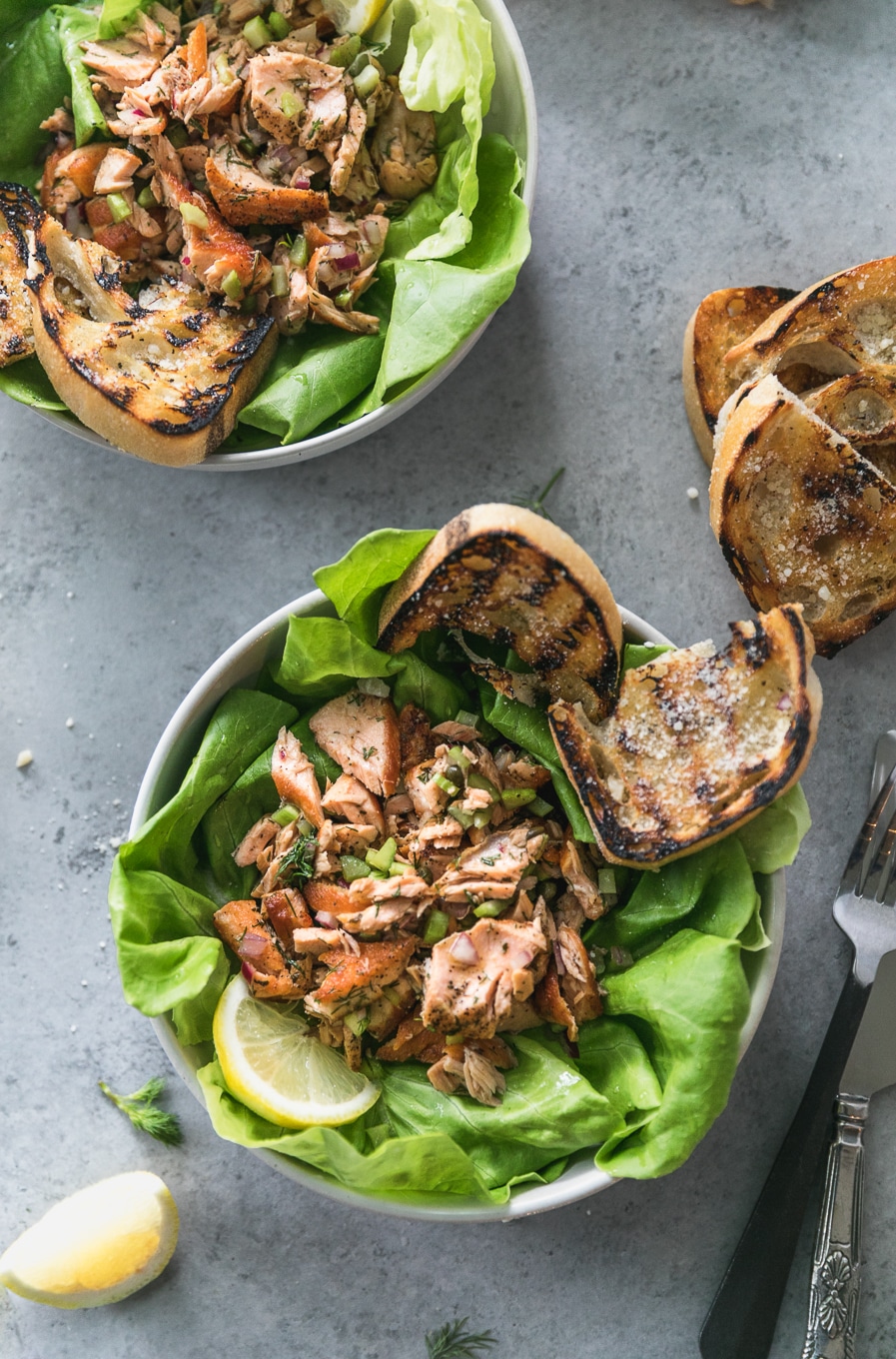 Overhead shot of two bowls filled with butter lettuce, flaked salmon and grilled garlic bread, with a lemon wedge and more slices of grilled garlic bread off to the side