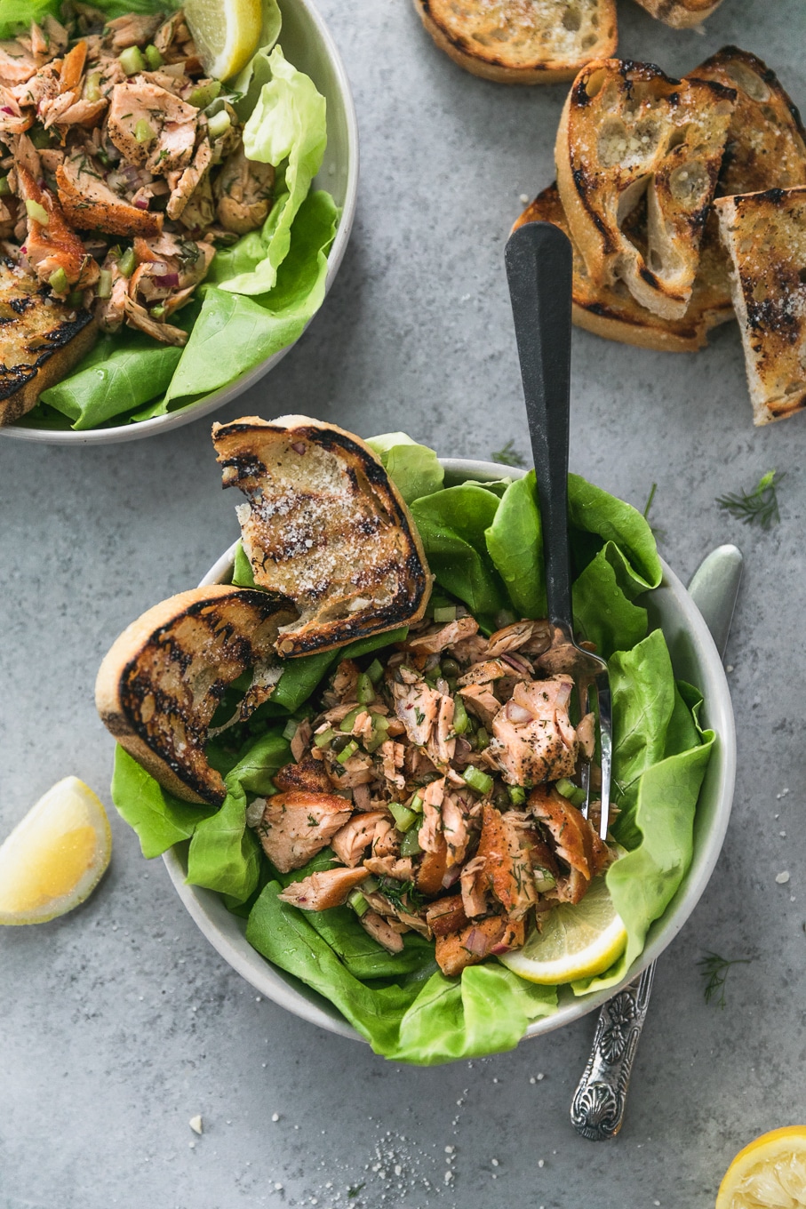 Overhead shot of two bowls filled with butter lettuce, flaked salmon and grilled garlic bread, with a lemon wedge and more slices of grilled garlic bread off to the side