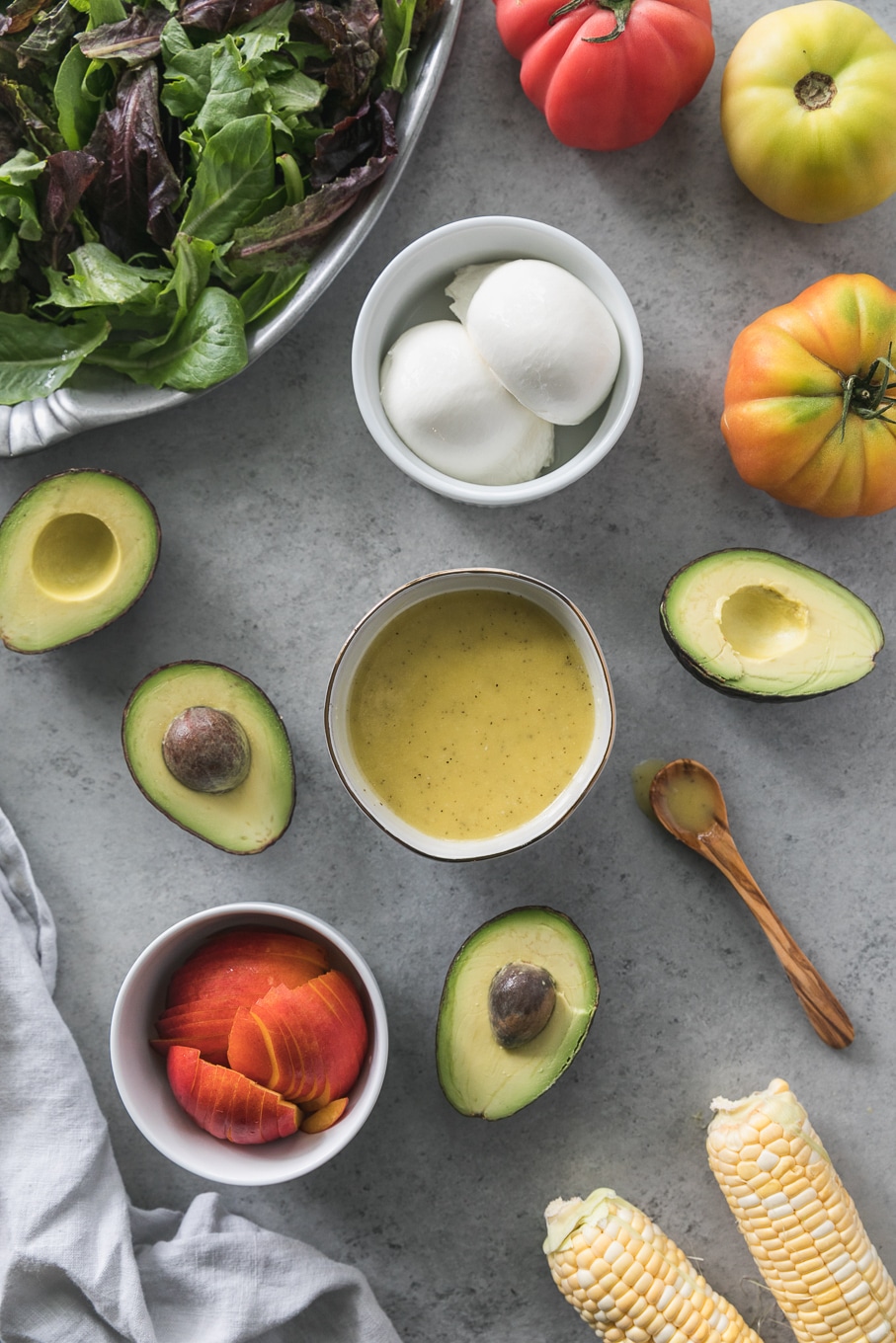 Overhead shot of a bowl of dressing surrounded by halved avocados, burrata, heirloom tomatoes, and sliced nectarines