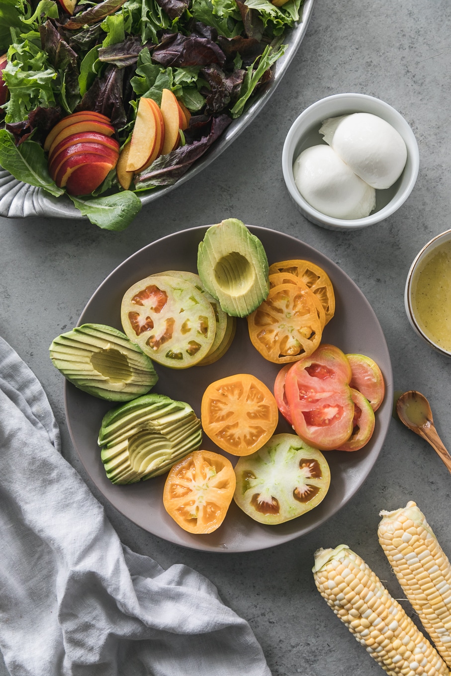 Overhead shot of a grey plate with sliced avocado and sliced heirloom tomatoes with corn on the cobb, burrata, and a bowl of dressing around it