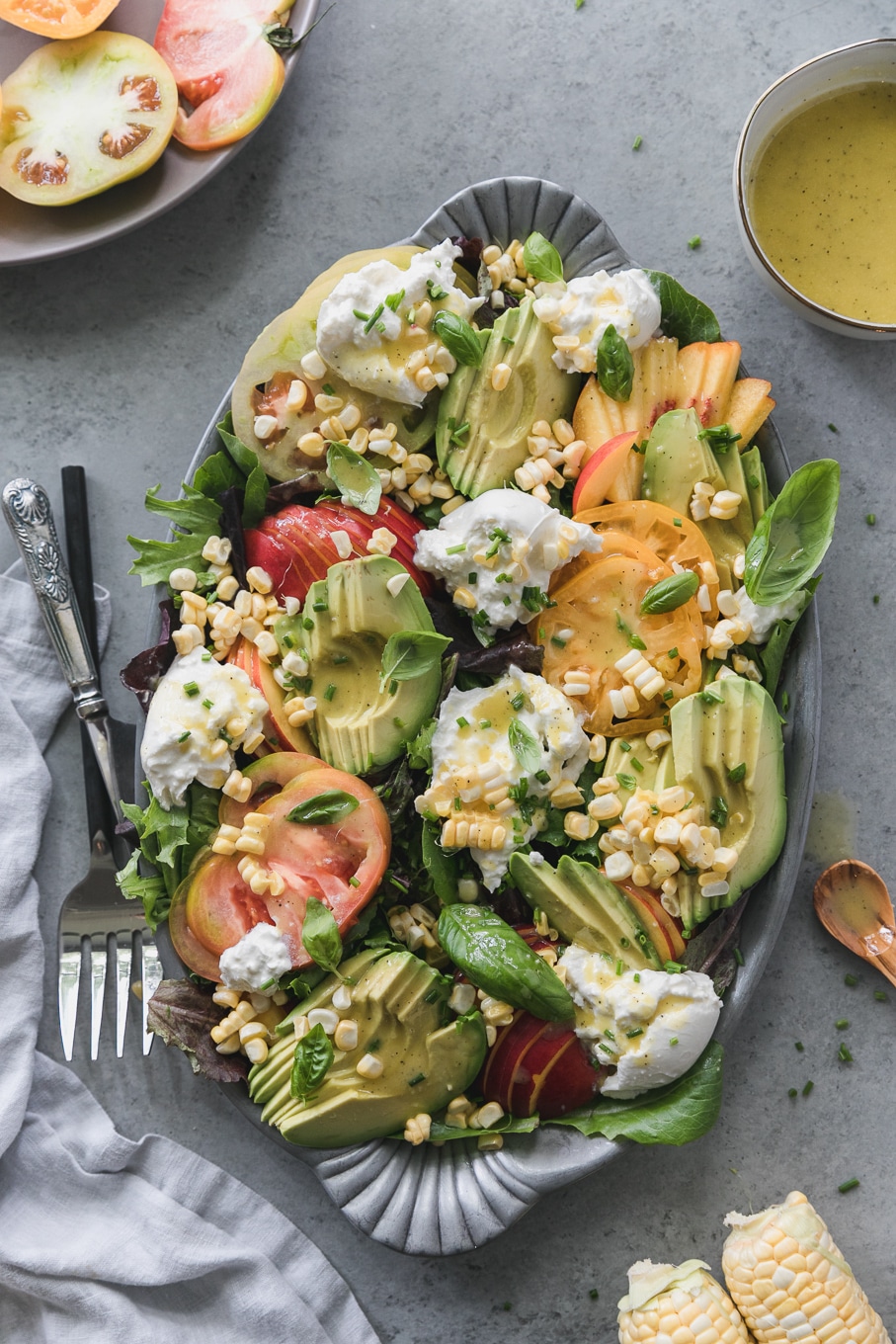Overhead shot of a salad on a platter topped with sliced avocado, nectarines, burrata, corn, heirloom tomatoes, and fresh herbs