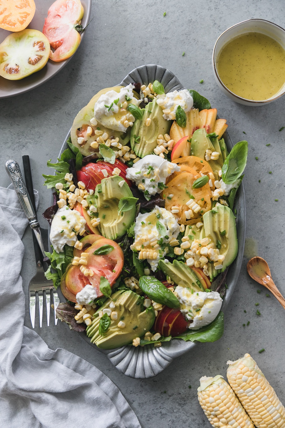 Overhead shot of a salad on a platter topped with sliced avocado, nectarines, burrata, corn, heirloom tomatoes, and fresh herbs