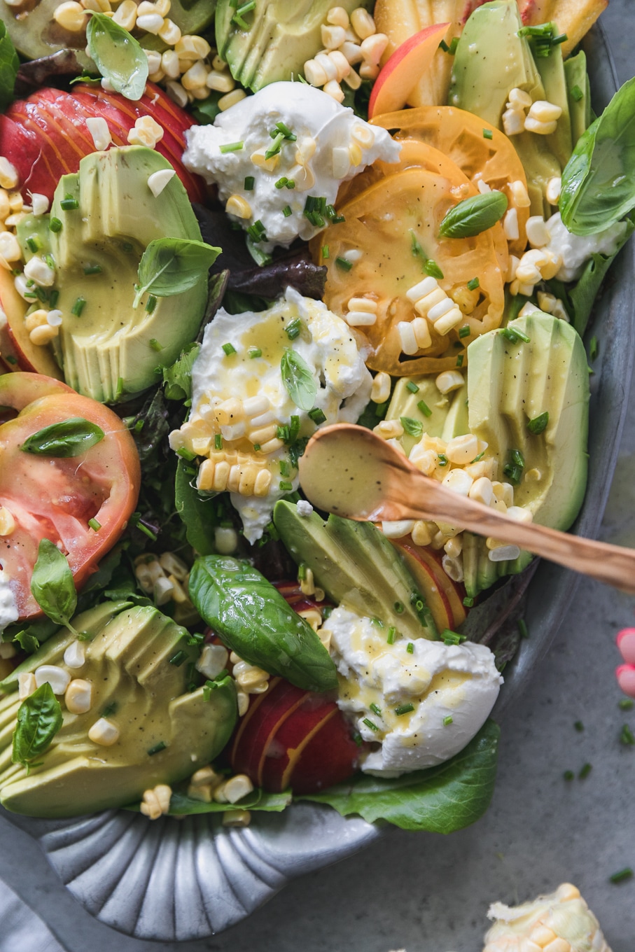 Overhead close up shot of an avocado, nectarine, corn, tomato, and burrata salad being drizzled with dressing on a wooden spoon