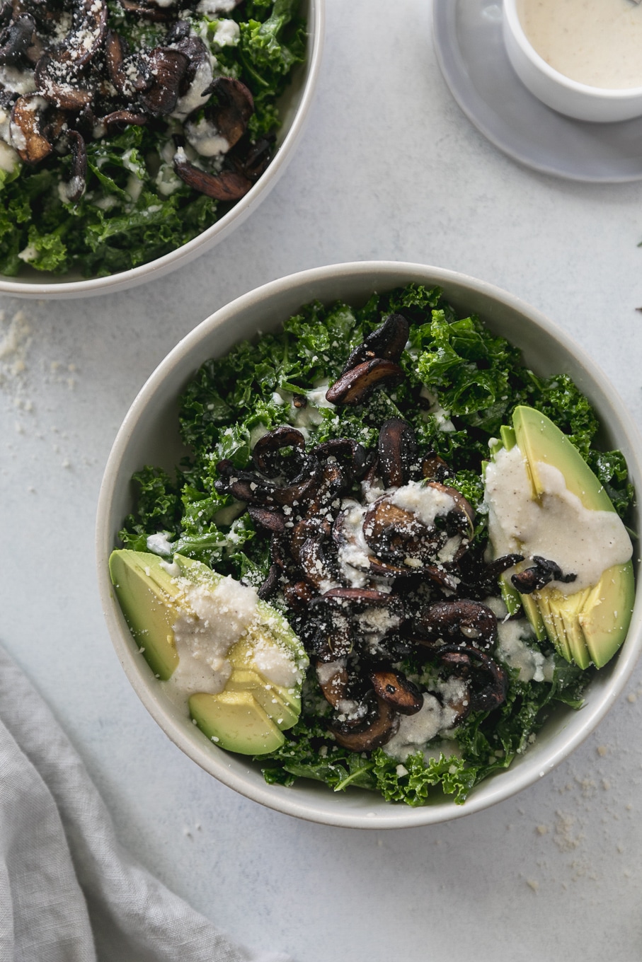 Overhead close up shot of a kale salad topped with mushrooms, creamy parmesan dressing, and sliced avocado with another kale salad sticking out of the left corner of the frame