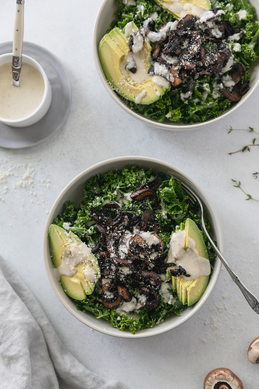 Overhead shot of two kale salads topped with mushrooms, sliced avocado, and creamy parmesan dressing with a bowl of dressing off to the side