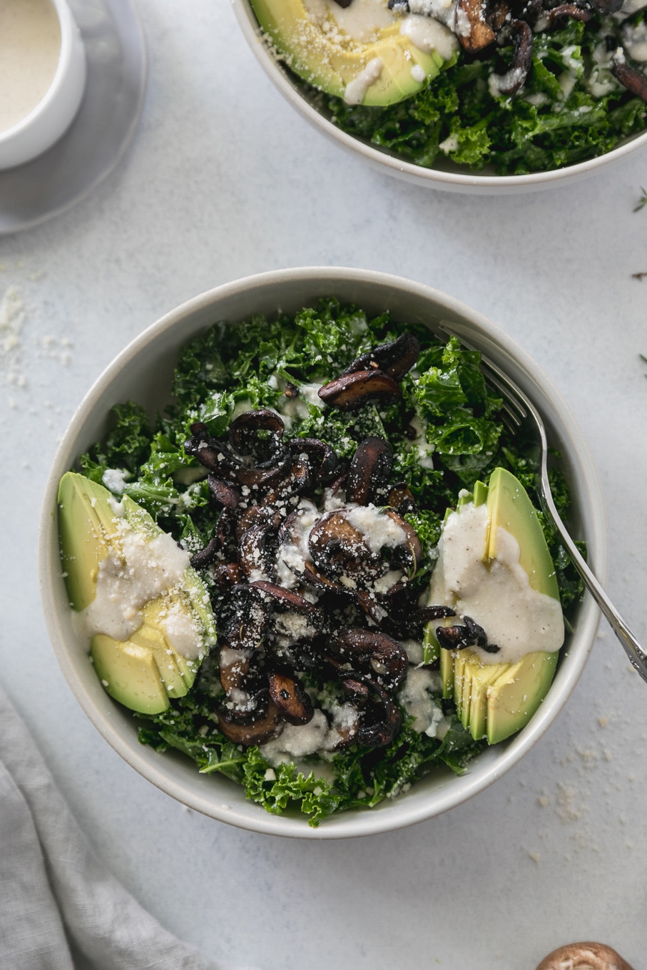Overhead shot of a kale salad topped with mushrooms, creamy parmesan dressing, and sliced avocado with a gold fork sticking out of the bowl