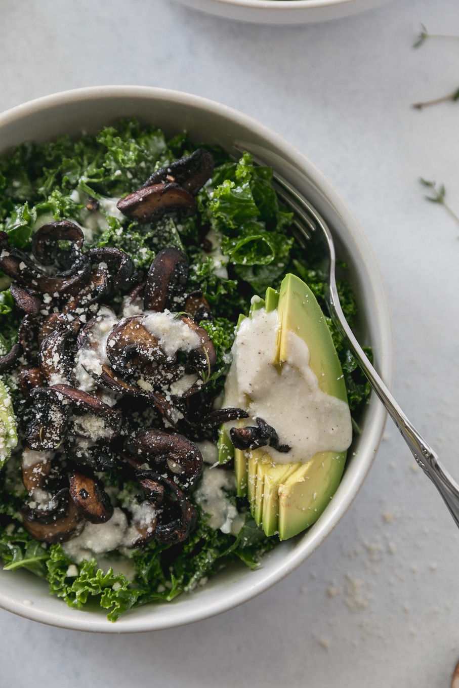 Overhead close up shot of a kale salad topped with mushrooms, creamy parmesan dressing, and sliced avocado with a gold fork sticking out of the bowl