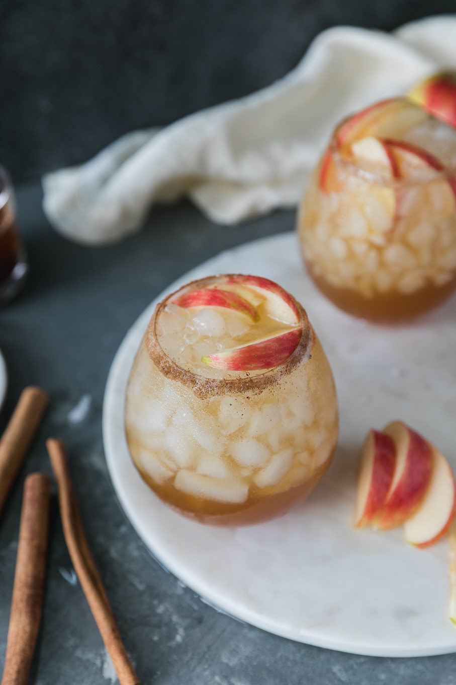 Close up shot of an apple pie bourbon fizz cocktail with another in the background along with sliced apples and cinnamon sticks and a cream colored linen