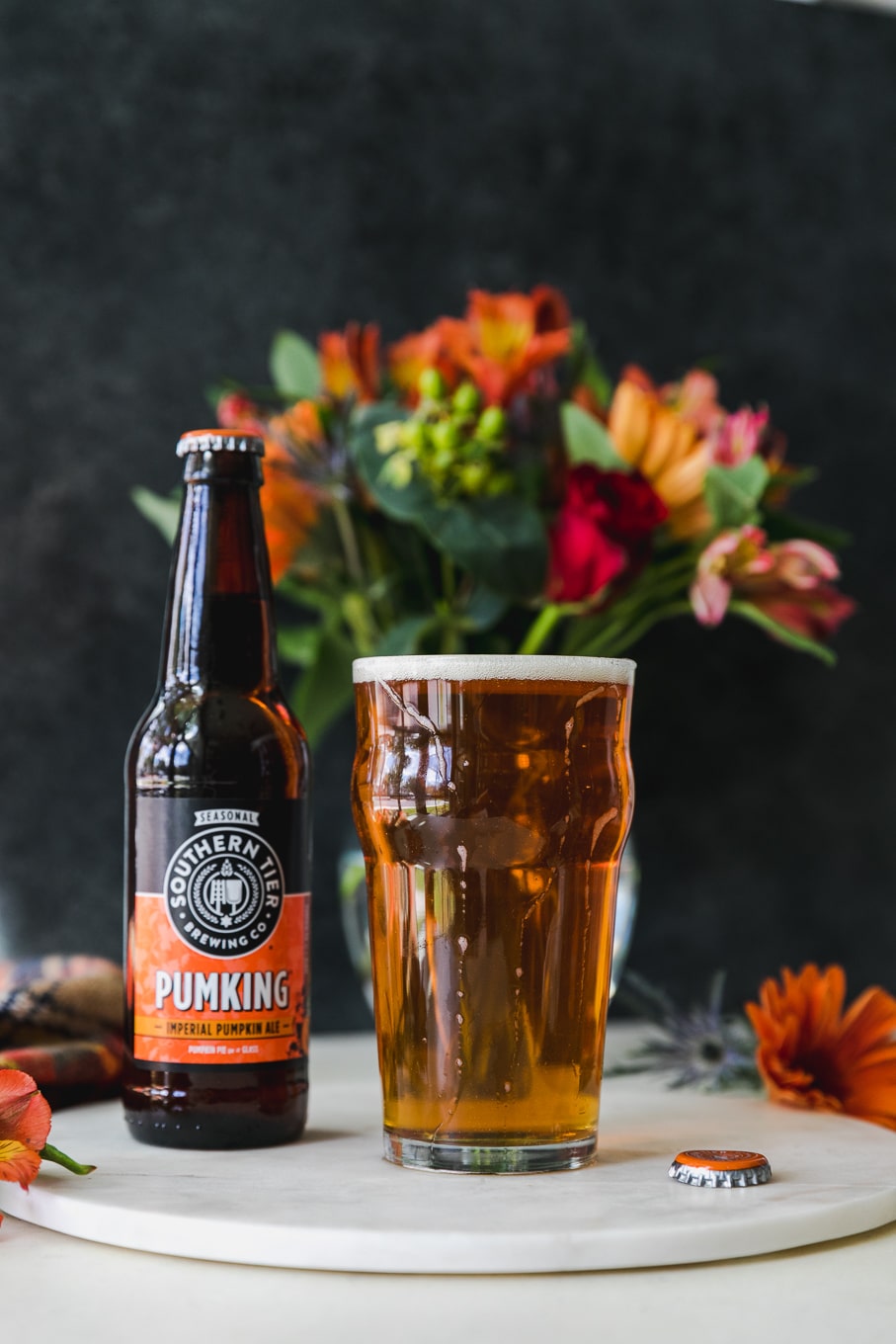 Forward facing shot of a beer glass filled with beer and a beer bottle next to it with a vase of flowers in the background and orange flowers scattered around