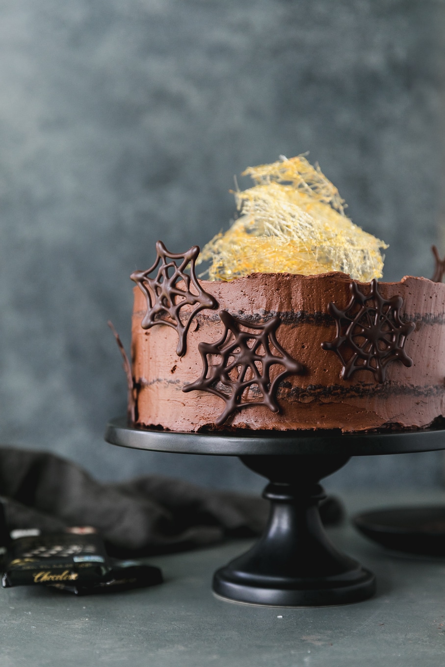 Forward facing shot of a chocolate cake with dark chocolate spider webs on it and a spun sugar cobweb on top on a black cake stand