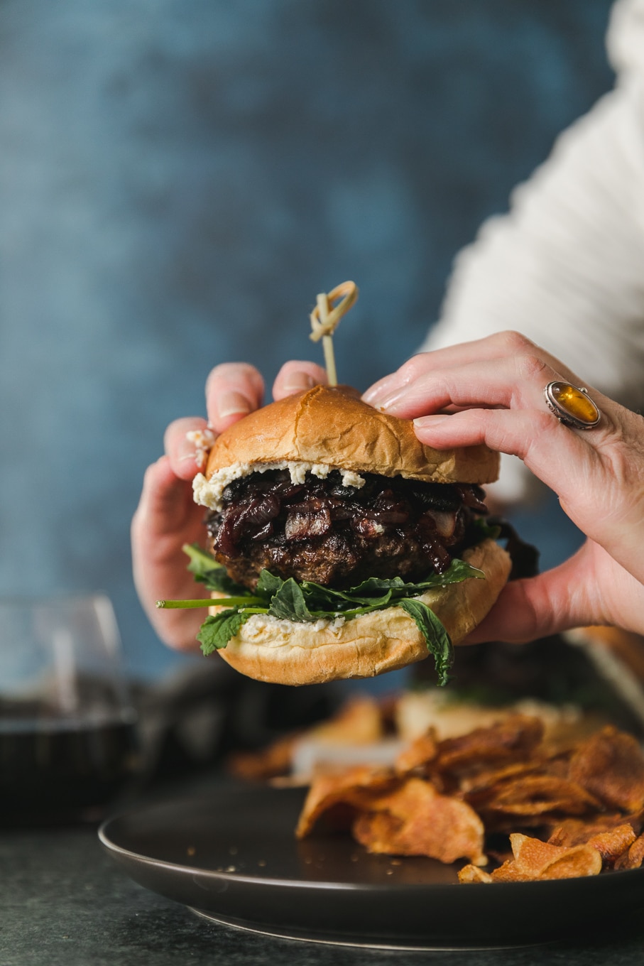 Close up forward facing shot of hands lifting up a burger from a plate of potato chips