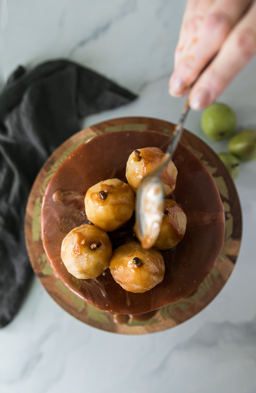 Overhead shot of a cake on a wooden and gold pedestal topped with poached pears, being drizzled with caramel sauce