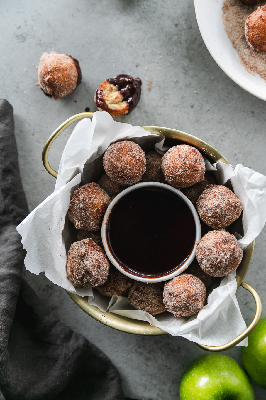 Overhead shot of a gold bowl with handles filled with doughnut holes and a ramekin of dark chocolate fudge sauce