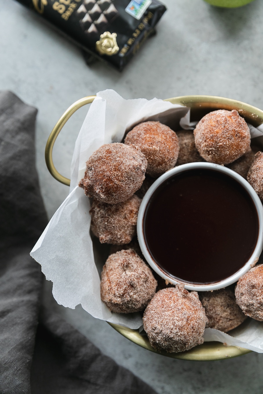 Overhead close up shot of a gold bowl with handles filled with doughnut holes and a ramekin of dark chocolate fudge sauce