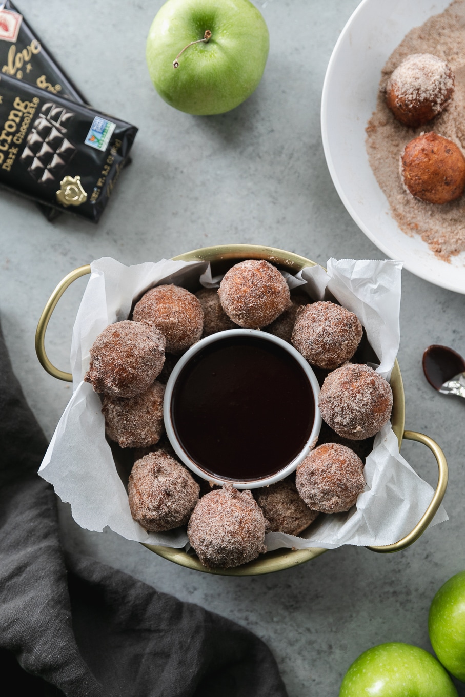 Overhead shot of a gold bowl with handles filled with doughnut holes and a ramekin of dark chocolate fudge sauce and chocolate bars above it