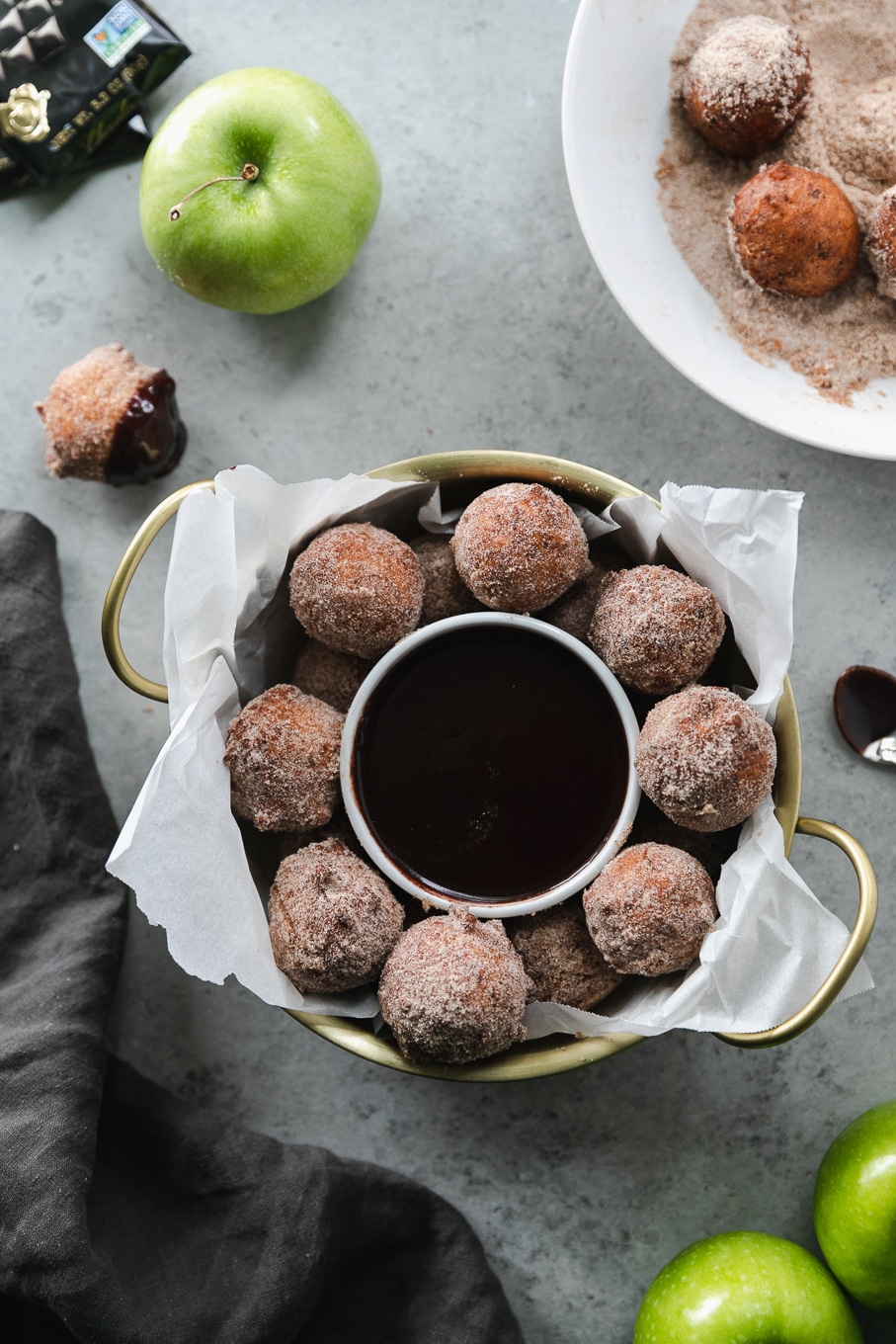 Overhead shot of a gold bowl with handles filled with doughnut holes and a ramekin of dark chocolate fudge sauce and a green apple above it