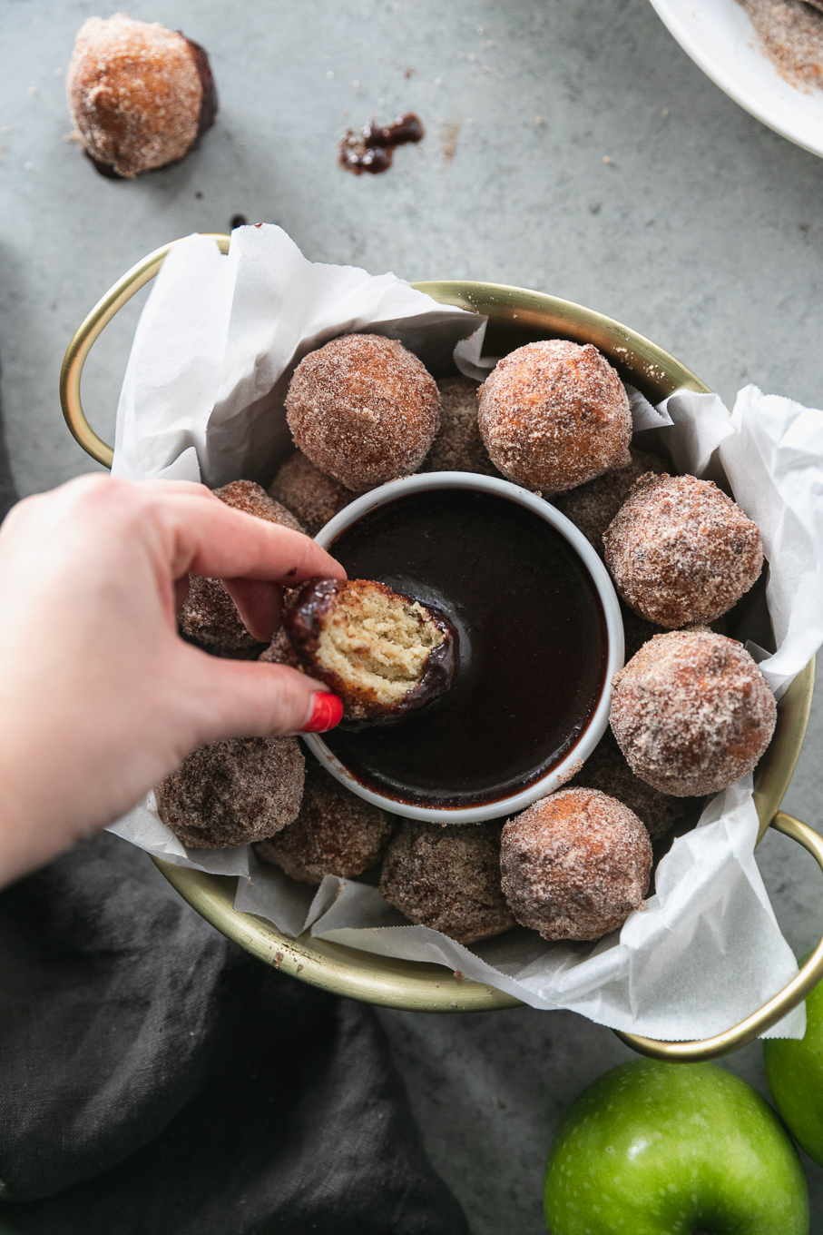 Overhead close up shot of a gold bowl with handles filled with doughnut holes and a hand dipping a doughnut hole into dark chocolate fudge sauce