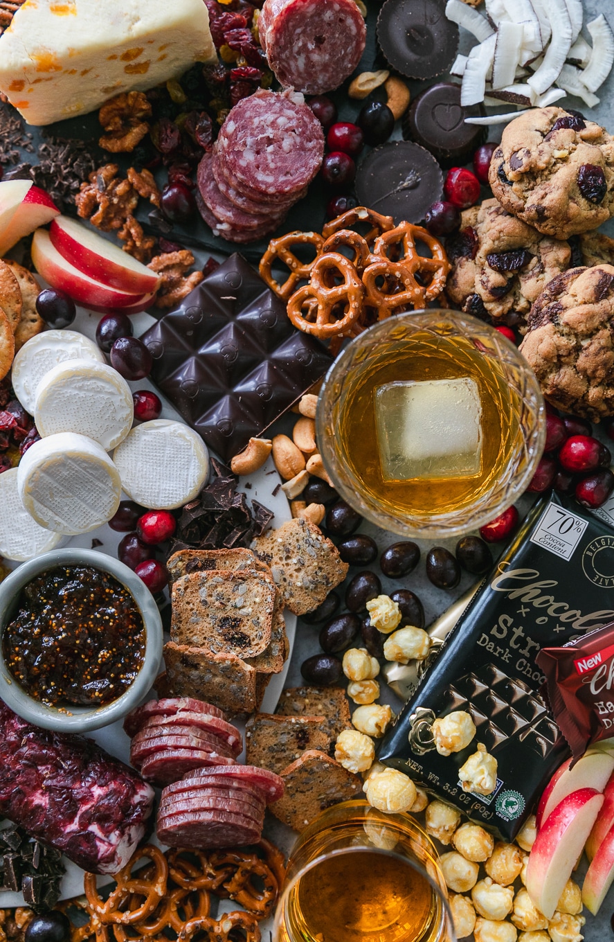Overhead shot of a chocolate and bourbon cheeseboard with apples, cranberries, crackers, pretzels, caramel corn, assorted cheeses, salami, and candy