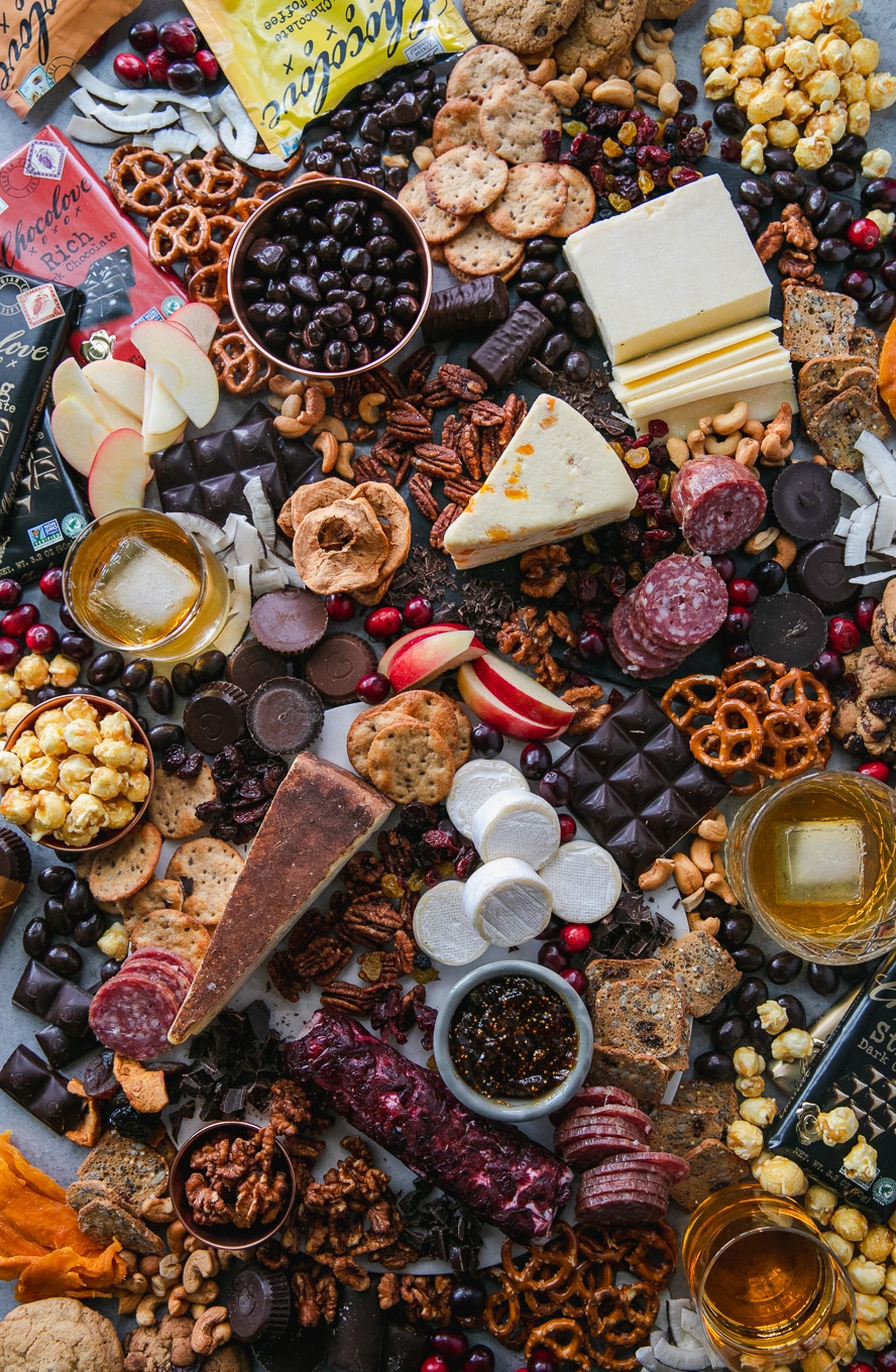 Overhead shot of a chocolate and bourbon cheeseboard with apples, cranberries, crackers, pretzels, caramel corn, assorted cheeses, salami, and candy