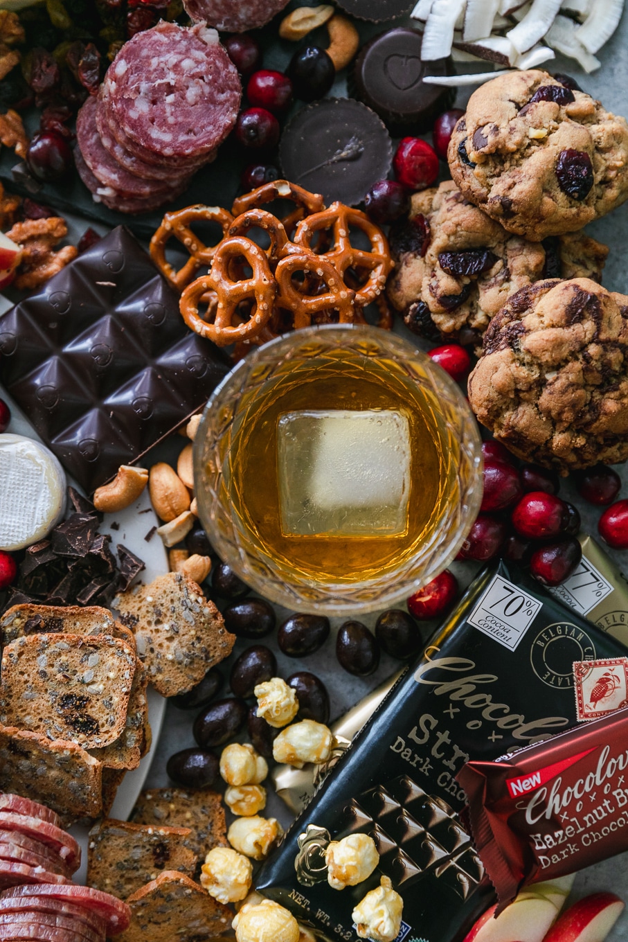 Overhead close up shot of a chocolate and bourbon cheeseboard with apples, cranberries, crackers, pretzels, caramel corn, assorted cheeses, salami, and candy