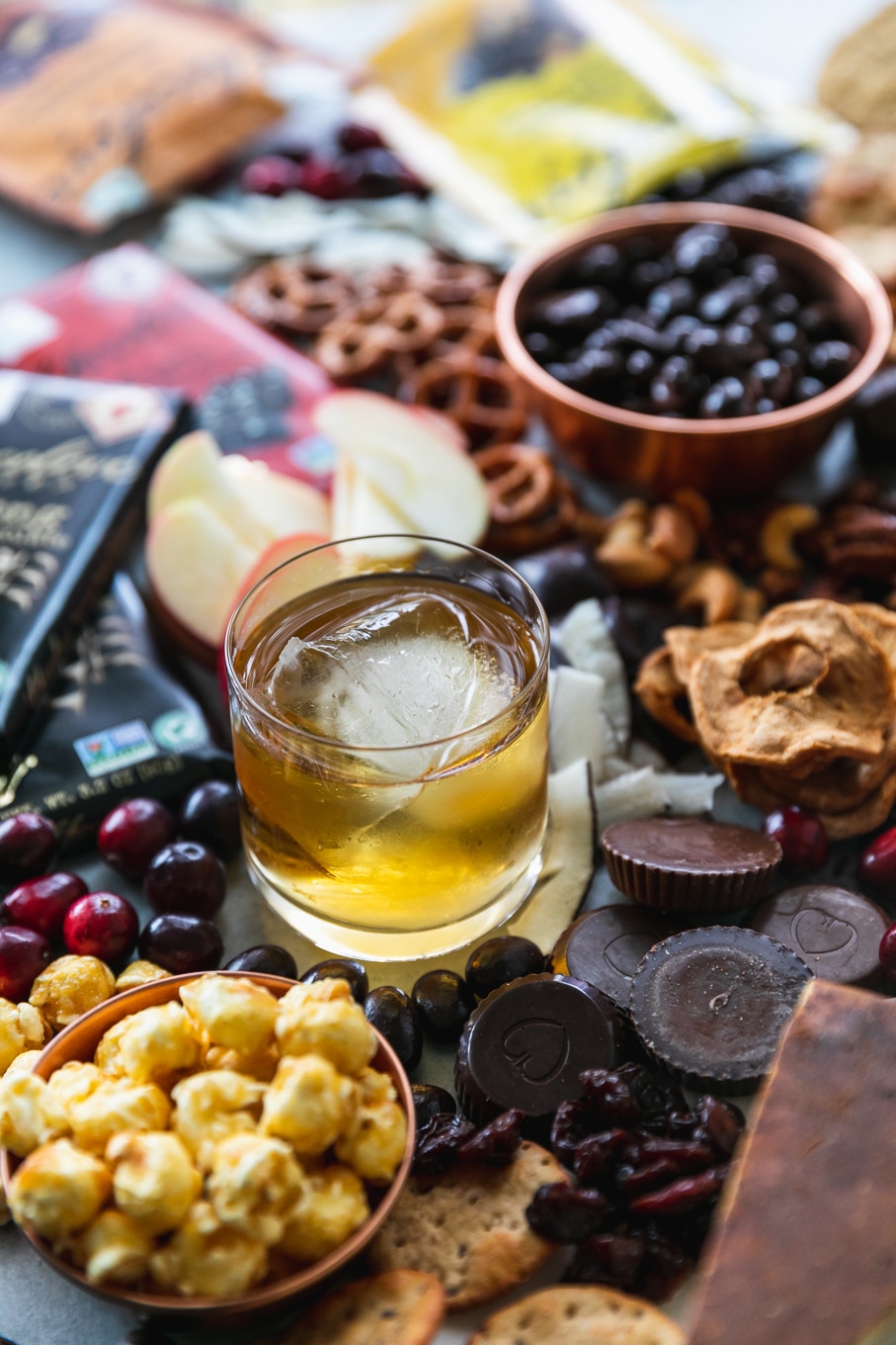 Close up shot of a glass of bourbon surrounded by cheeses,dried fruit, chocolate, pretzels, and caramel corn
