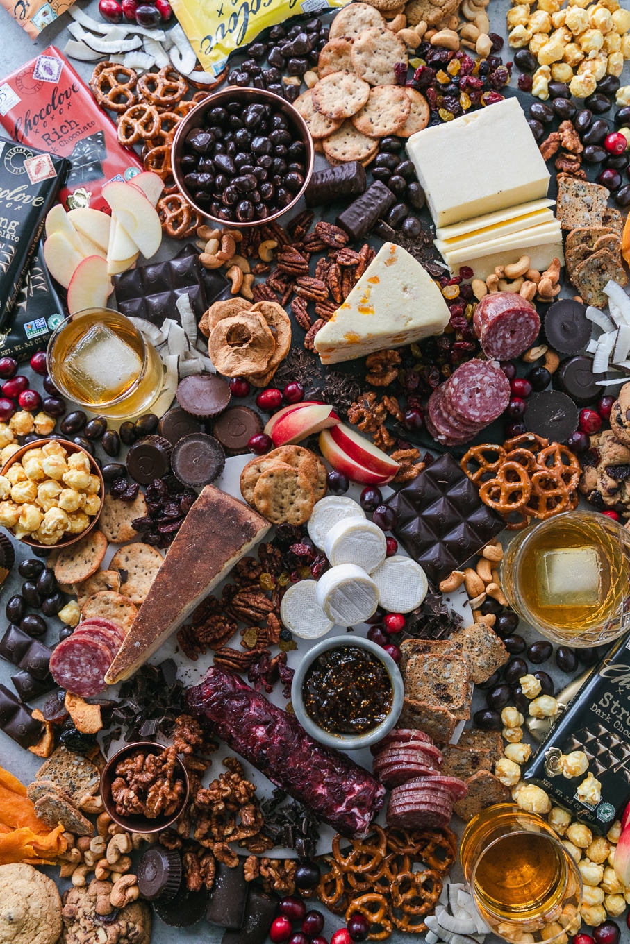 Overhead shot of a chocolate and bourbon cheeseboard with apples, cranberries, crackers, pretzels, caramel corn, assorted cheeses, salami, and candy