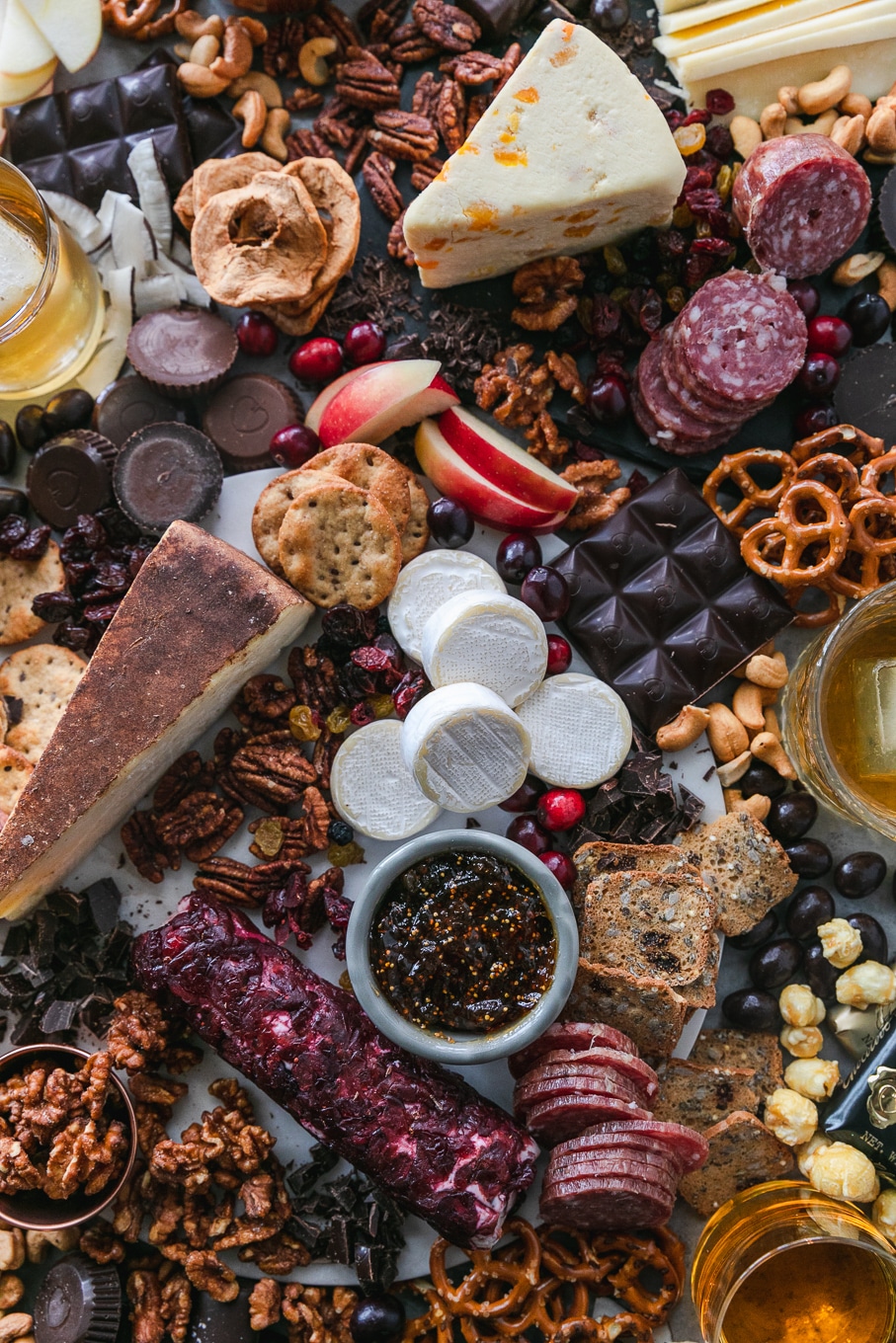 Overhead shot of a chocolate and bourbon cheeseboard with apples, cranberries, crackers, pretzels, caramel corn, assorted cheeses, salami, and candy