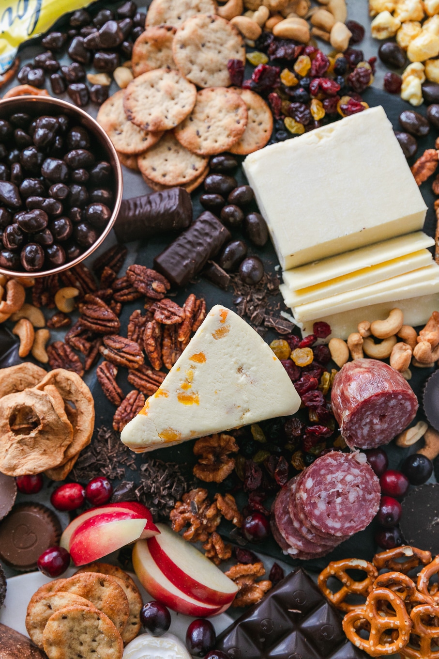 Overhead close up shot of a chocolate and bourbon cheeseboard with apples, cranberries, crackers, pretzels, caramel corn, assorted cheeses, salami, and candy