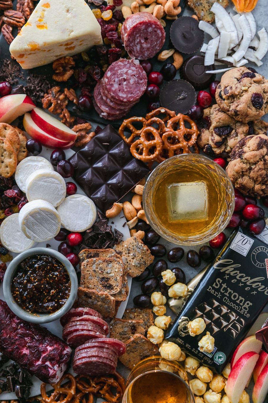 Overhead shot of a chocolate and bourbon cheeseboard with apples, cranberries, crackers, pretzels, caramel corn, assorted cheeses, salami, and candy