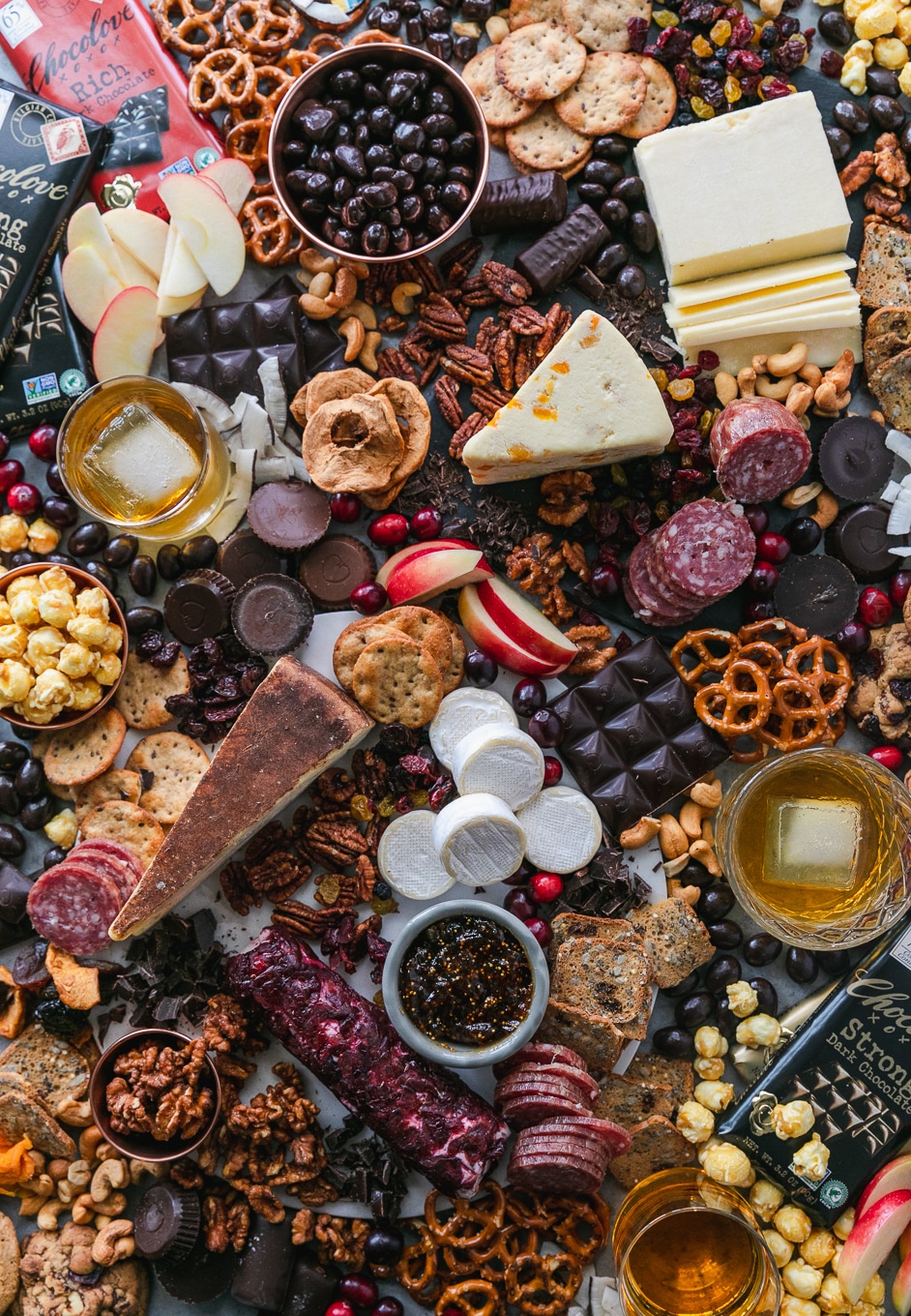 Overhead shot of a chocolate and bourbon cheeseboard with apples, cranberries, crackers, pretzels, caramel corn, assorted cheeses, salami, and candy