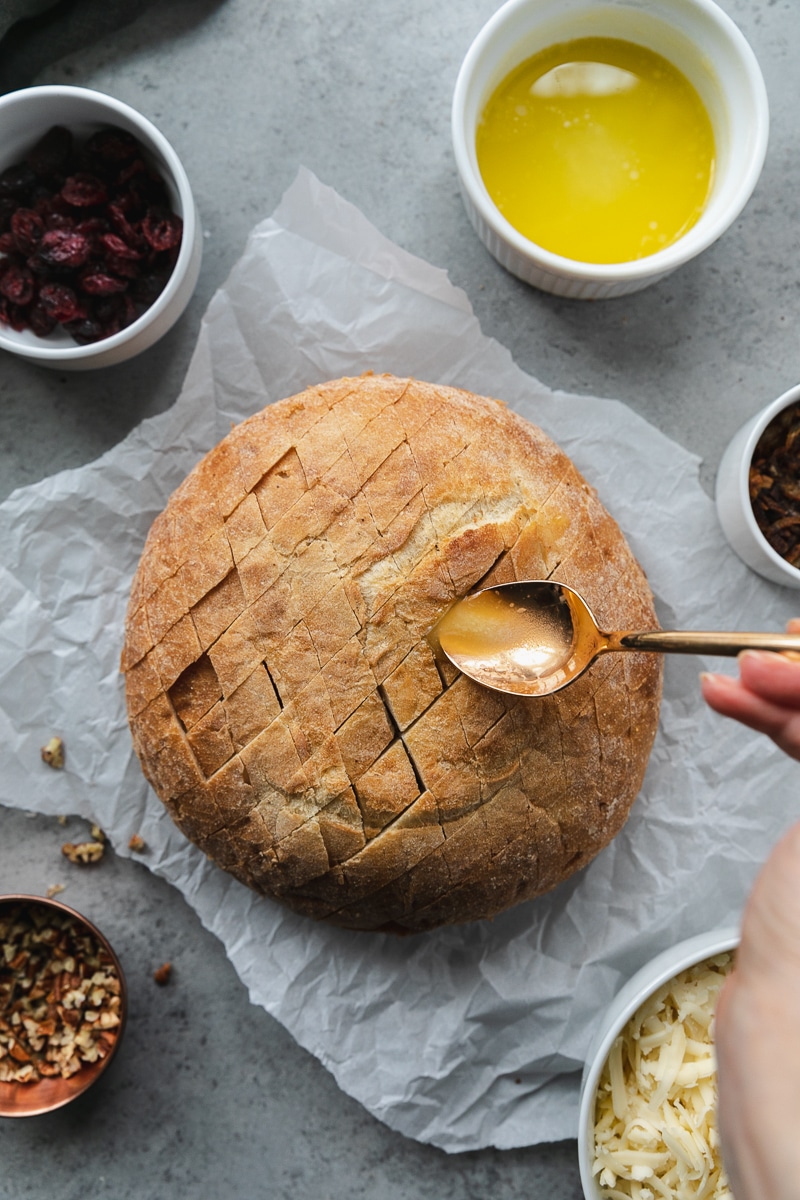 Overhead shot of a loaf of bread with diagonal cuts in it, with a gold spoon drizzling melted butter on it