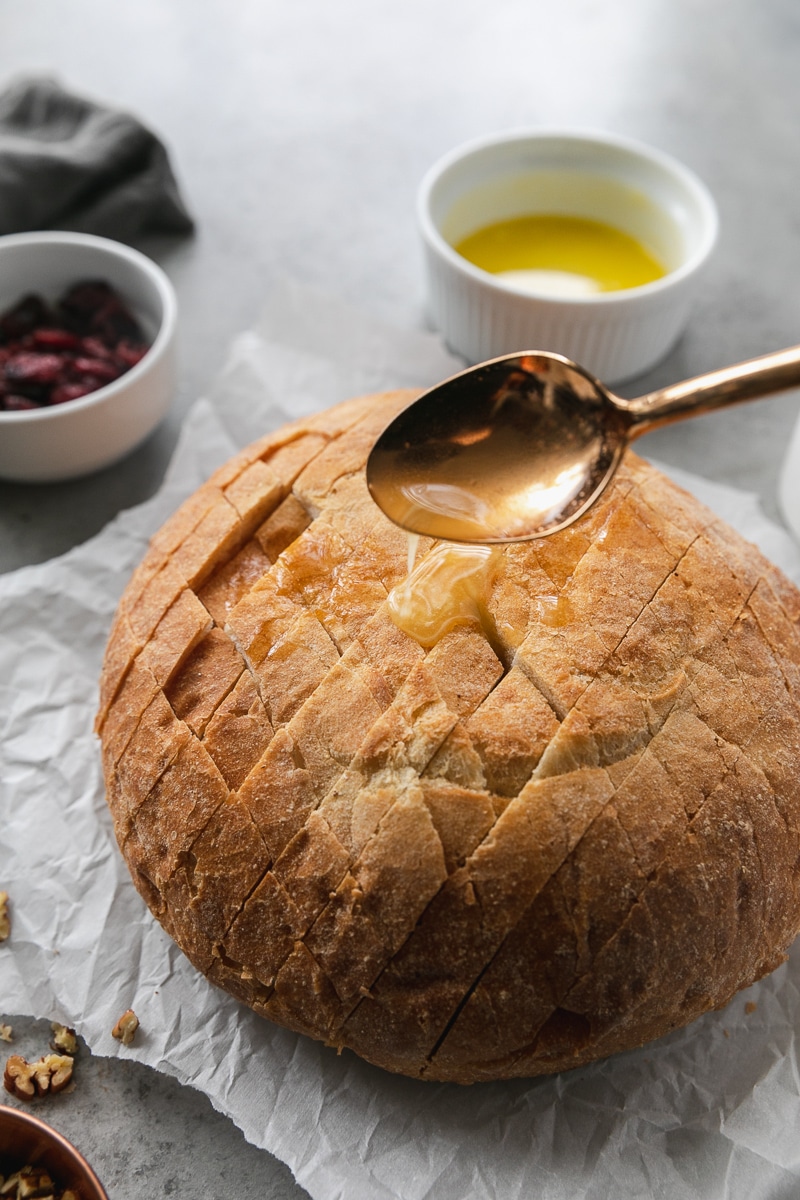 Close up shot of a loaf of bread with diagonal cuts in it, with a gold spoon drizzling melted butter on it
