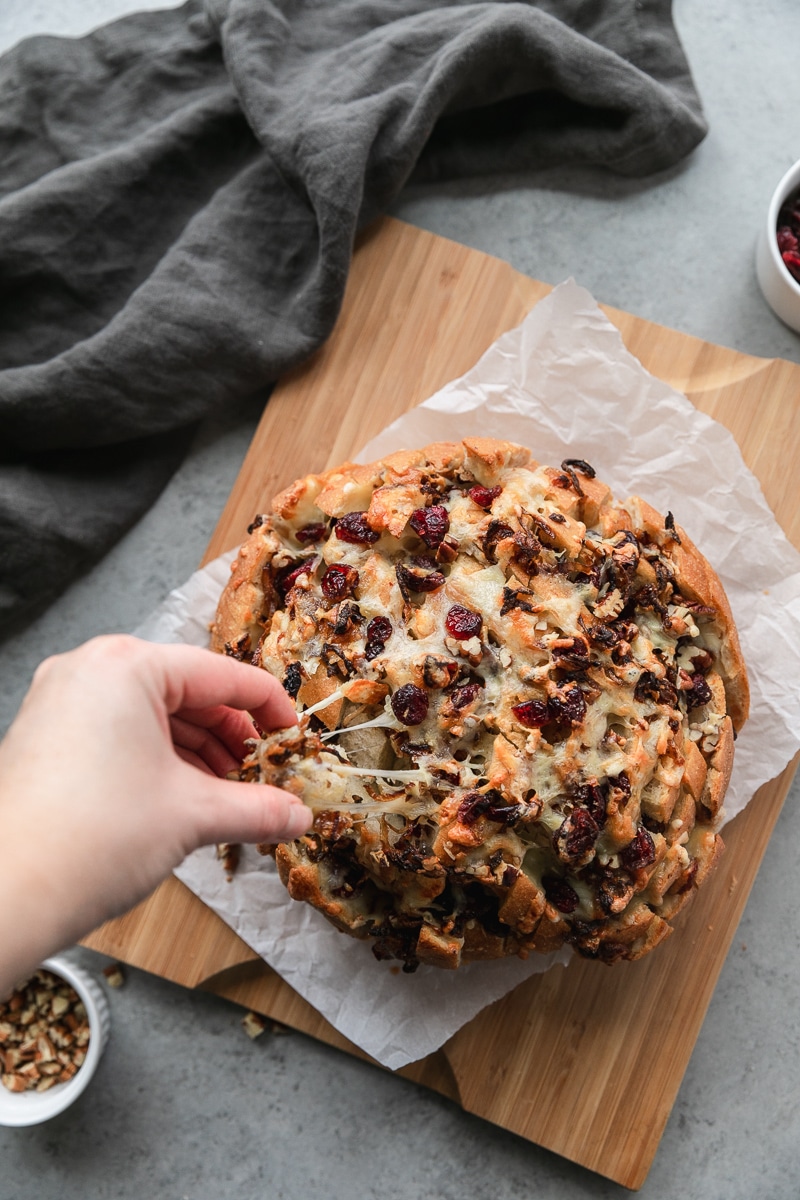 Overhead shot of a circular loaf of bread stuffed with melted cheese, caramelized onions, cranberries, and pecans with a hand pulling a cheesy piece of bread off of it
