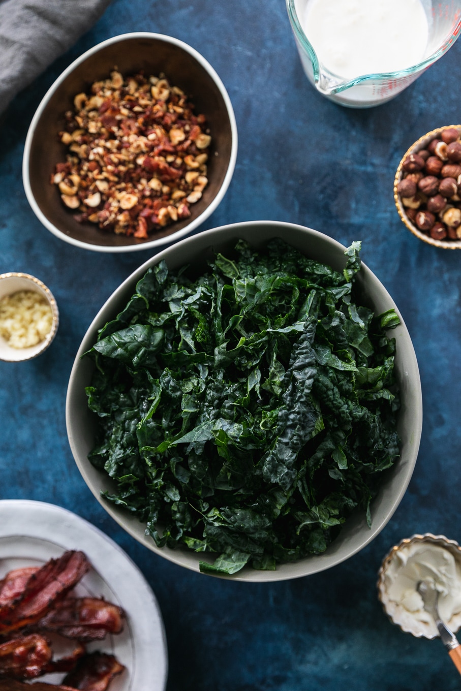 Overhead shot of a bowl of shredded kale, a bowl of chopped hazelnuts, a plate of bacon, a ramekin of garlic, and a ramekin of cream cheese on a dark blue background