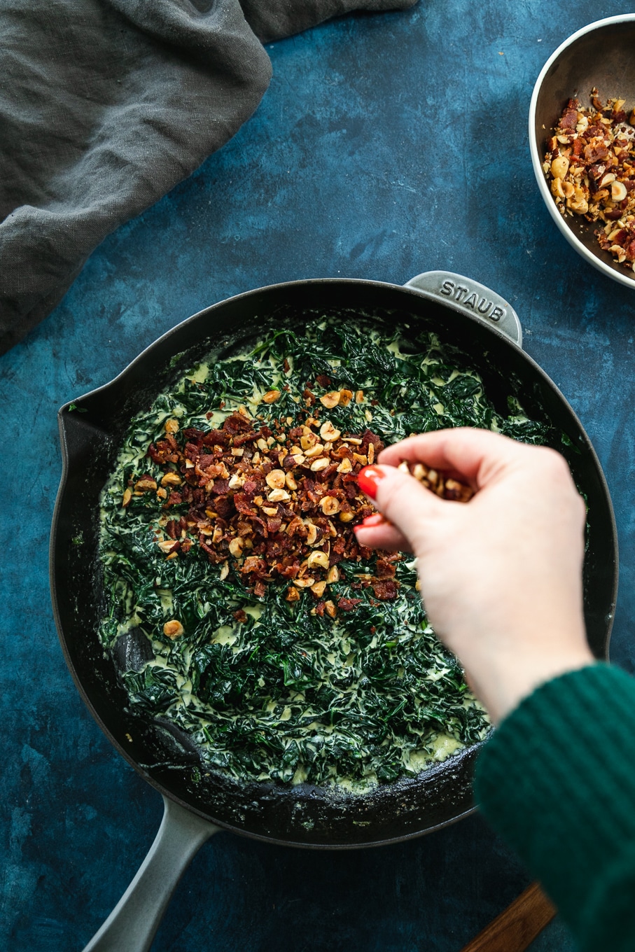 Overhead shot of a cast iron skillet filled with creamed kale and a hand sprinkling hazelnut bacon crunch on top